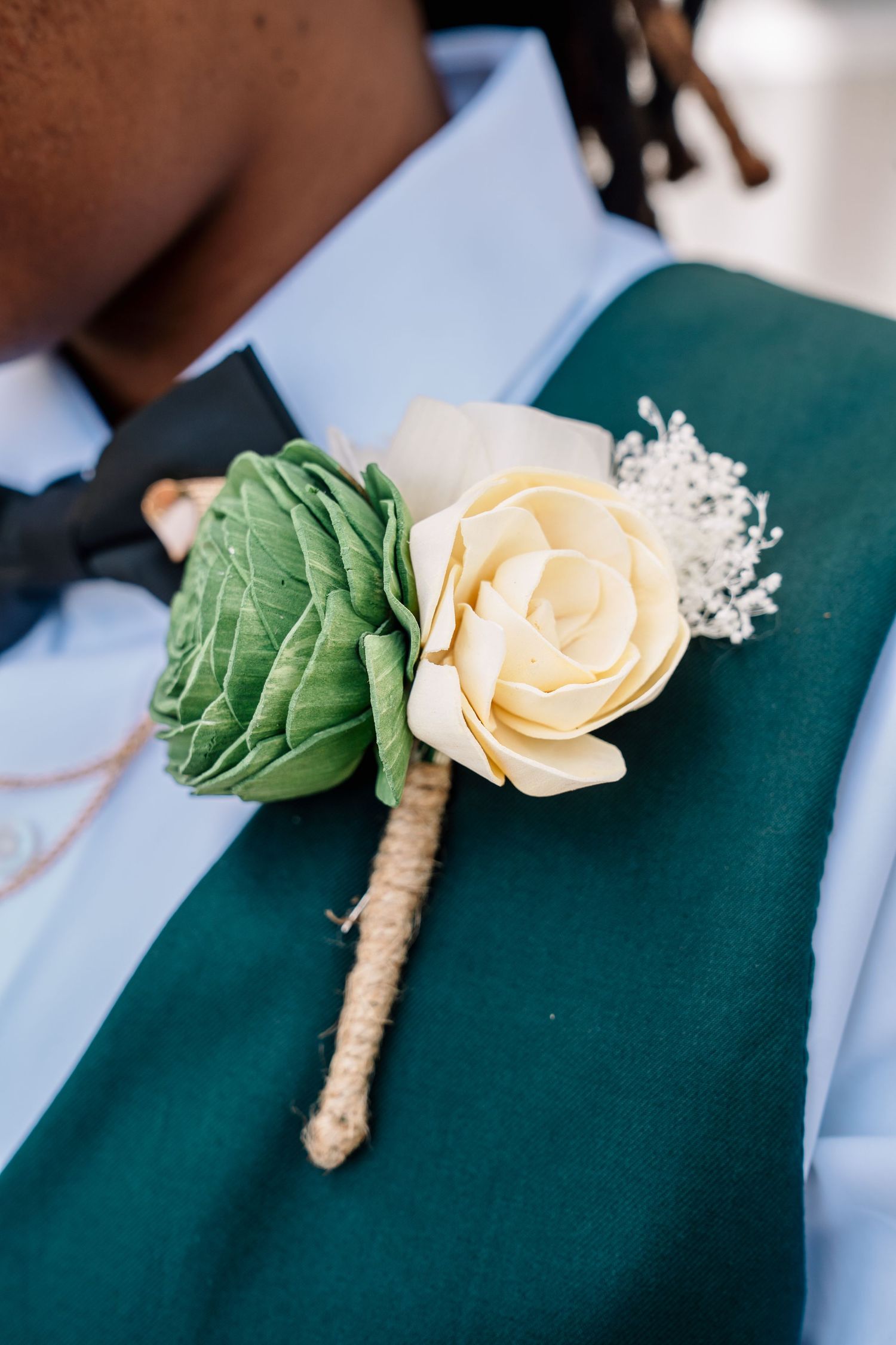 Close-up of a cream rose boutonniere with greenery and baby's breath on a teal wedding vest.