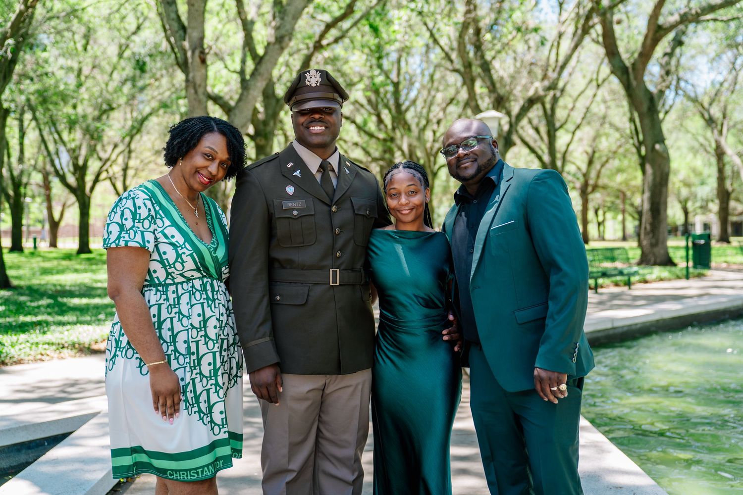 Family of four posing outdoors near a fountain, one member in military dress uniform, others in elegant green attire.