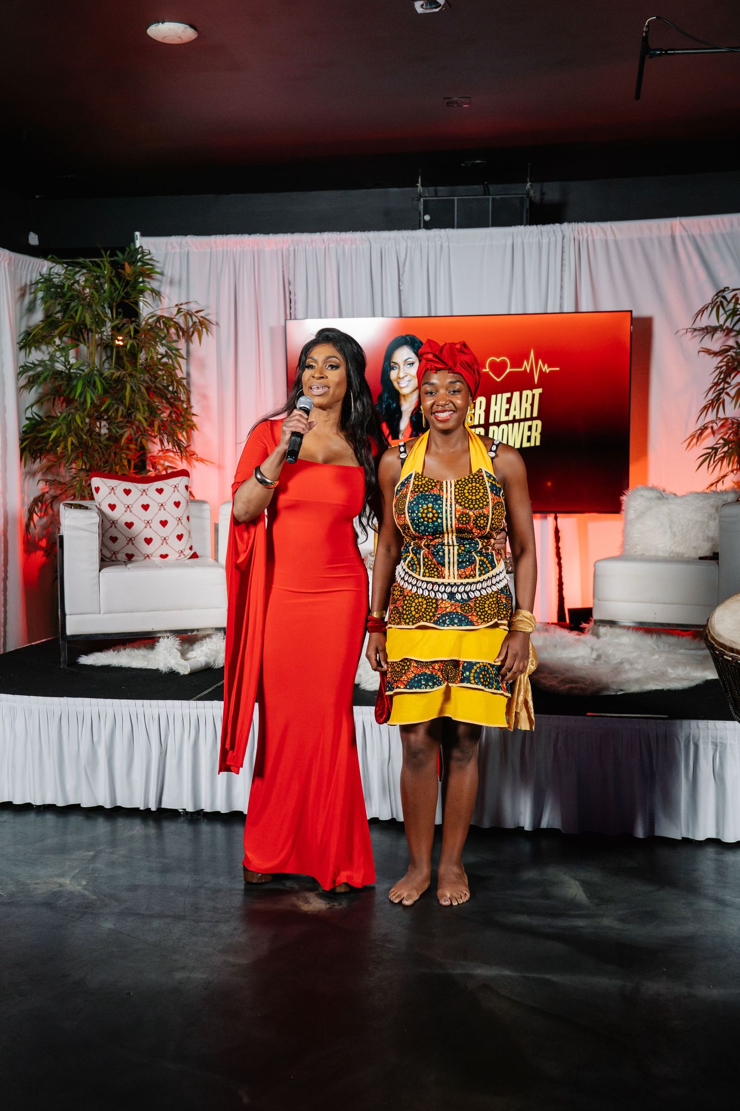 Two women posing at a Heart Power event, one in a red gown holding a mic and one in African print dress.