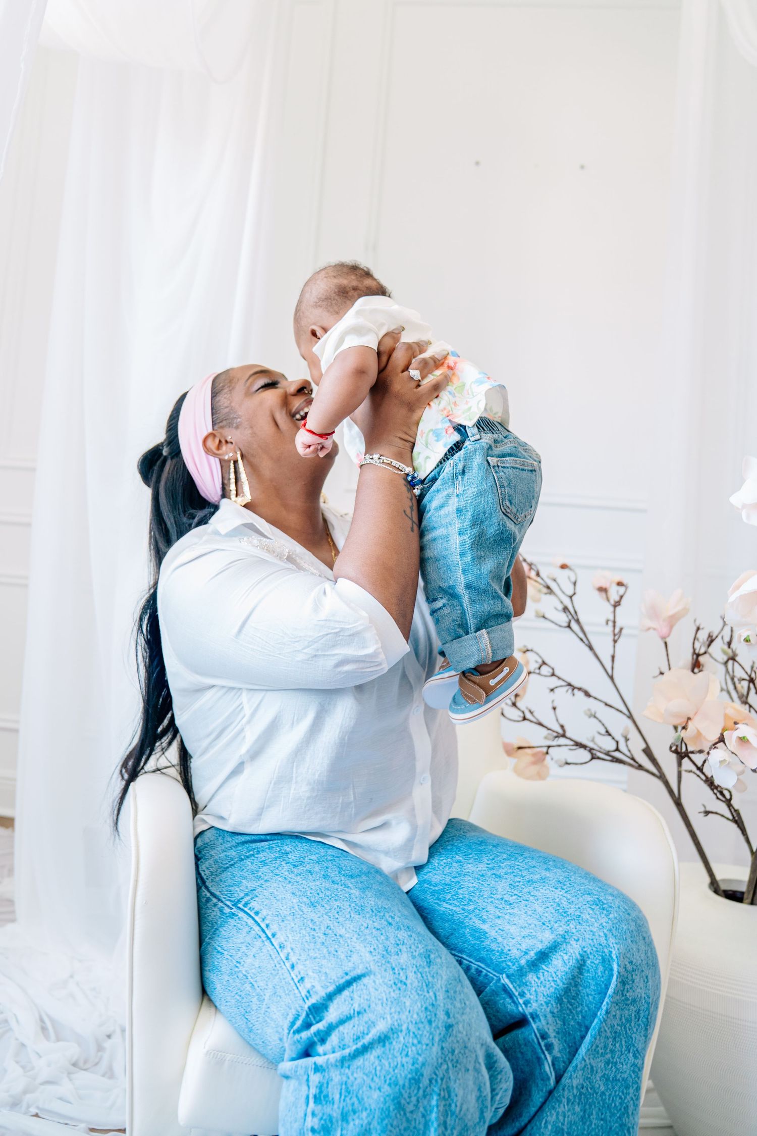 A mother sits in a white chair, joyfully lifting her baby up to her face in a bright, airy room.