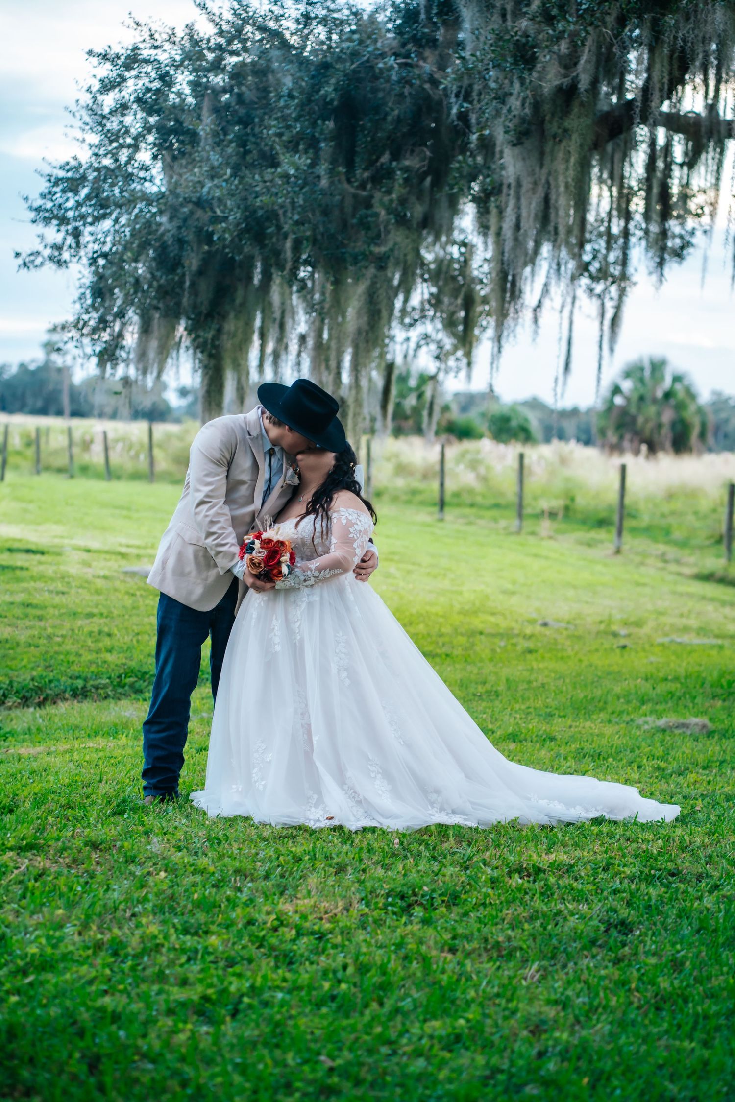 Couple in wedding attire share a romantic moment on a lush green lawn beneath Spanish moss draped trees.
