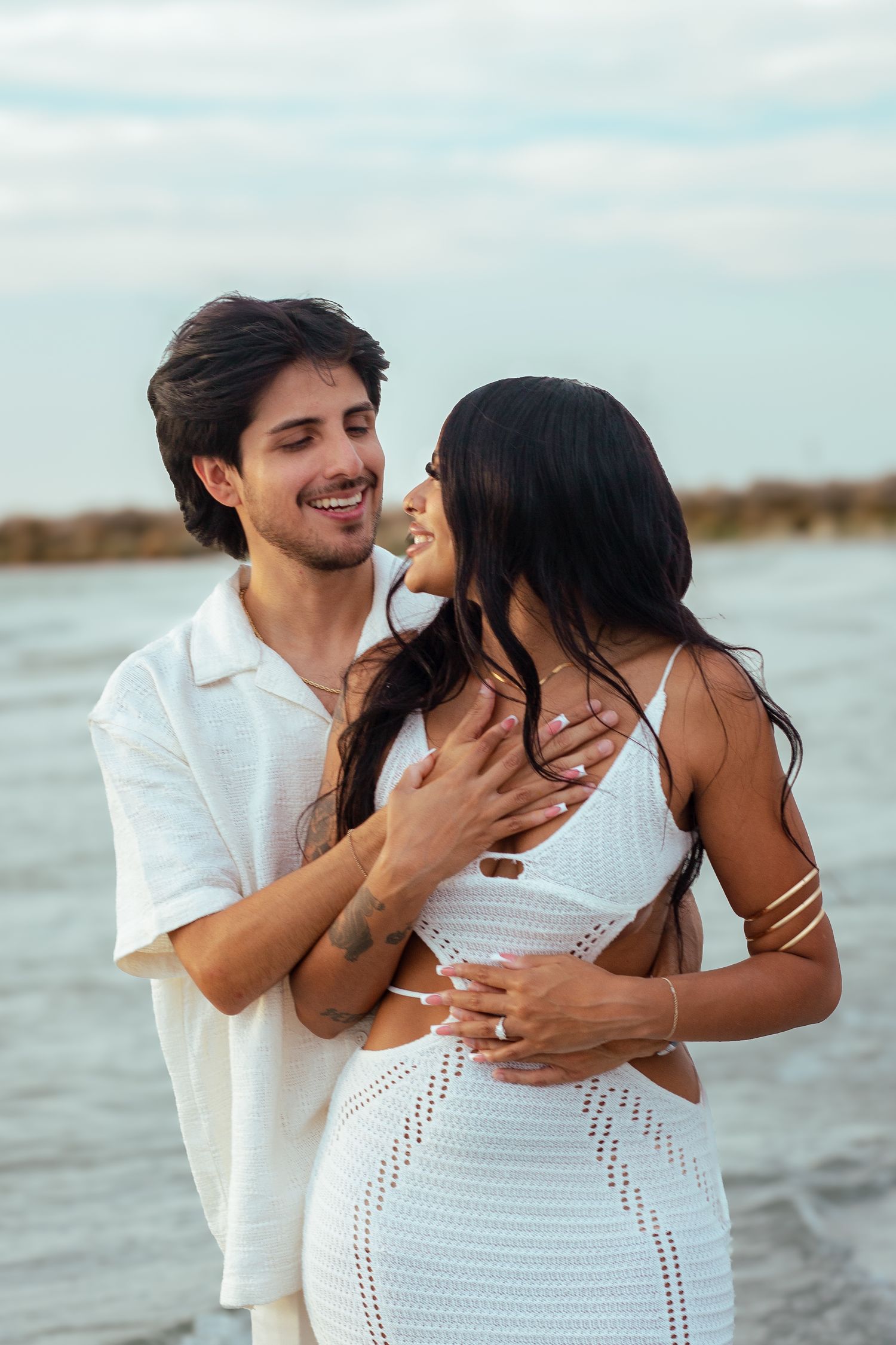 Affectionate beach photoshoot with couple in white outfits by the water.