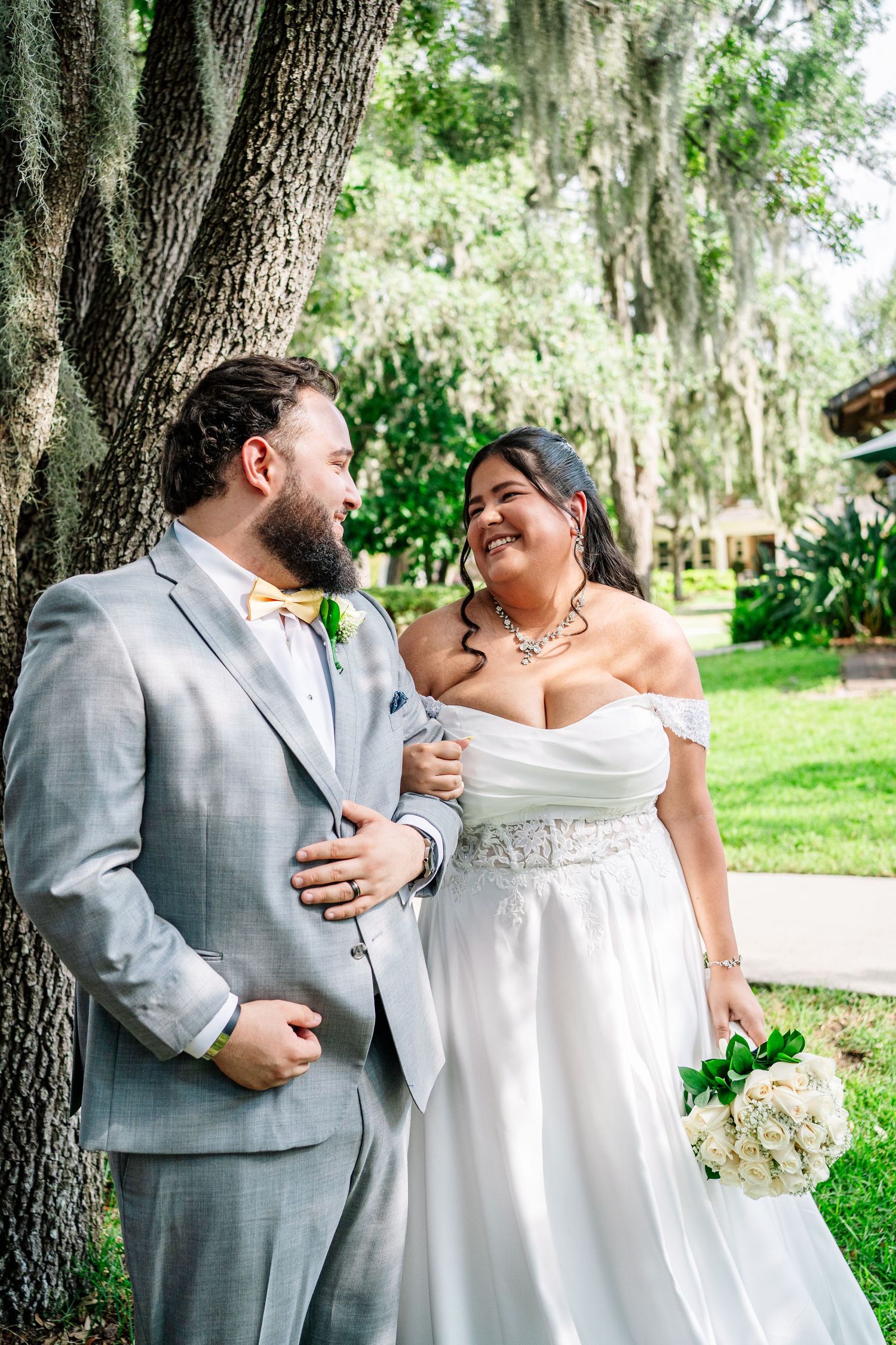 A smiling bride and groom in wedding attire stand together under a moss-covered tree in a lush outdoor setting.