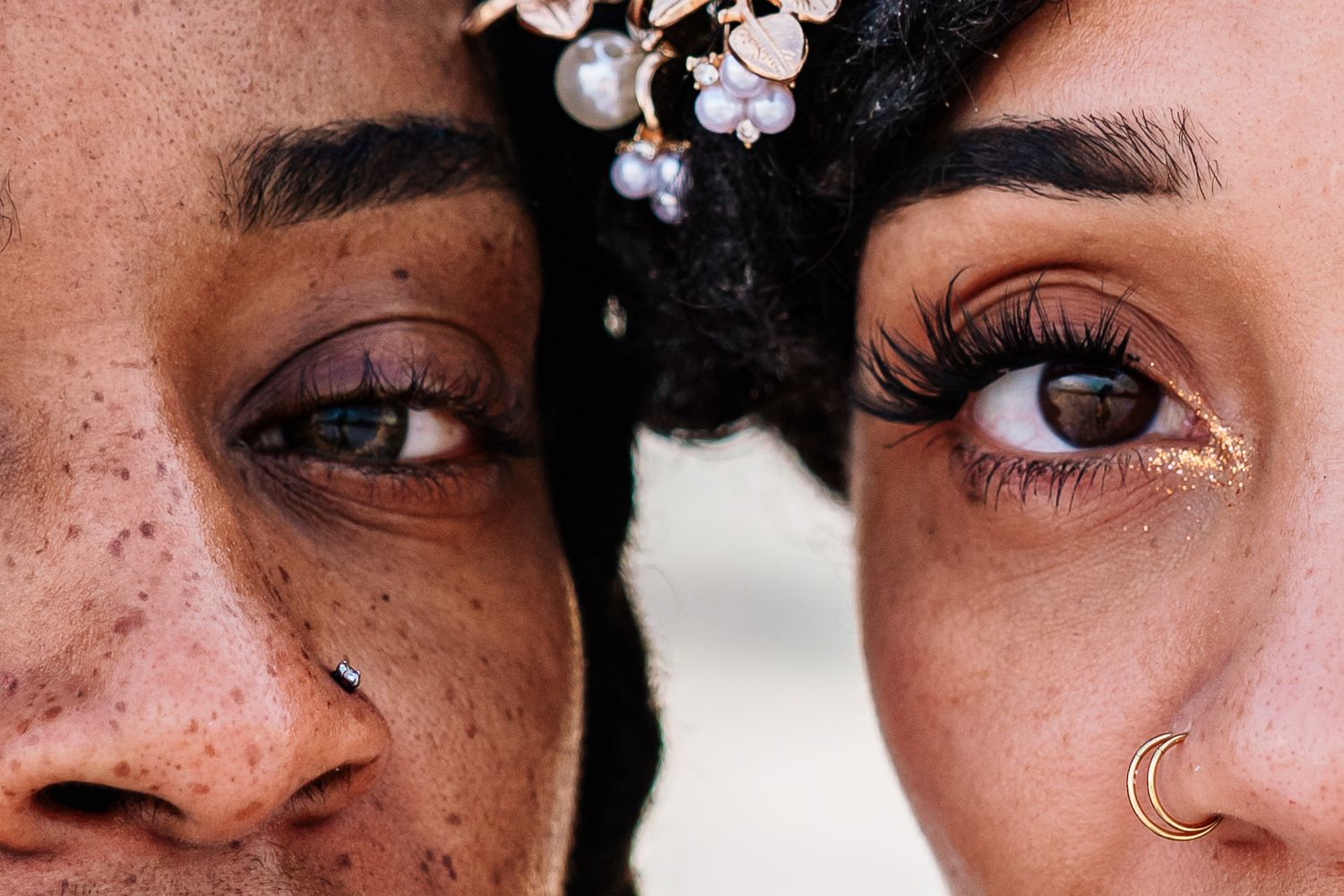 Close-up of two faces side by side, showcasing freckles, nose piercings, and dramatic eyelashes.