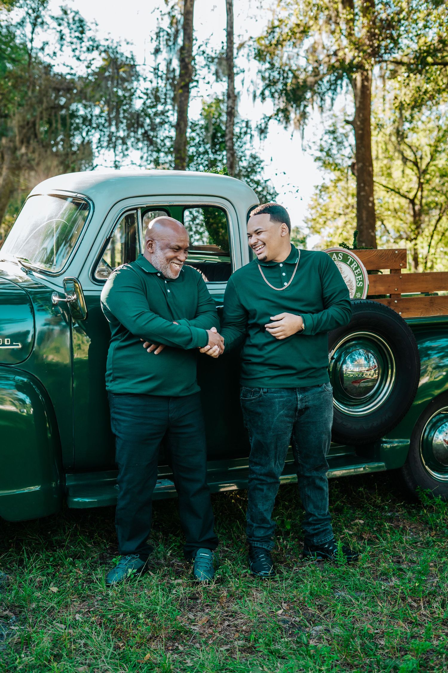 Two people in matching green sweaters share a laugh while standing by a classic pickup truck.