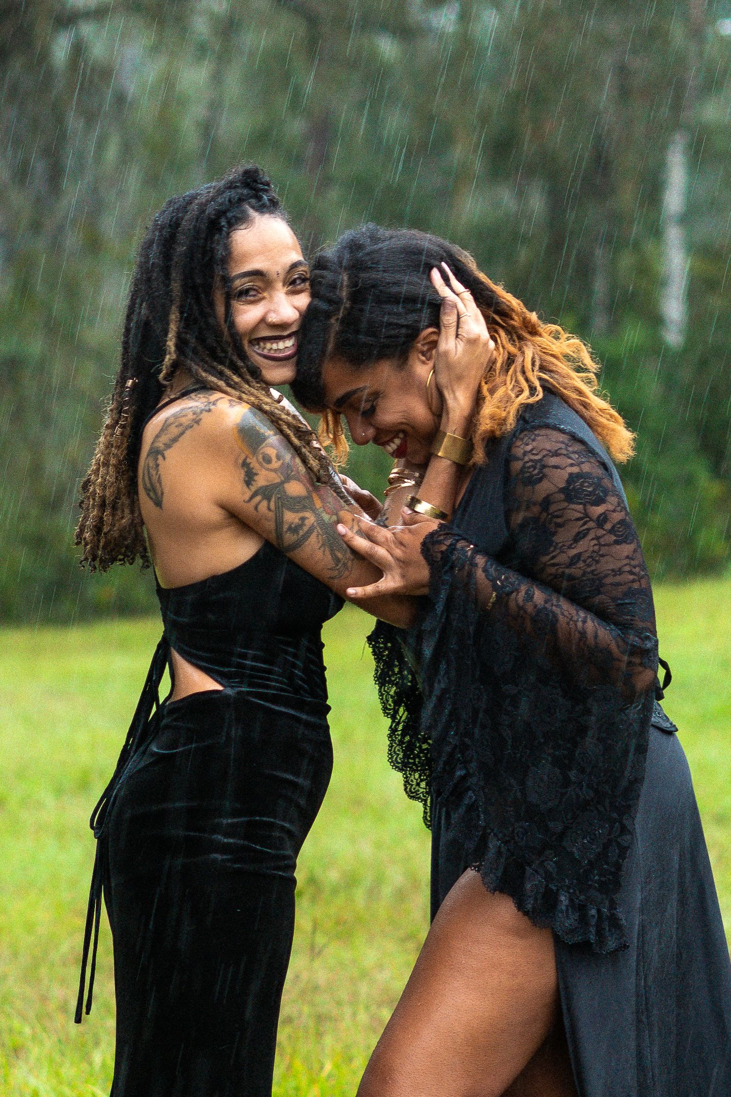 Two people in black outfits dance and laugh together in the rain on a grassy field.