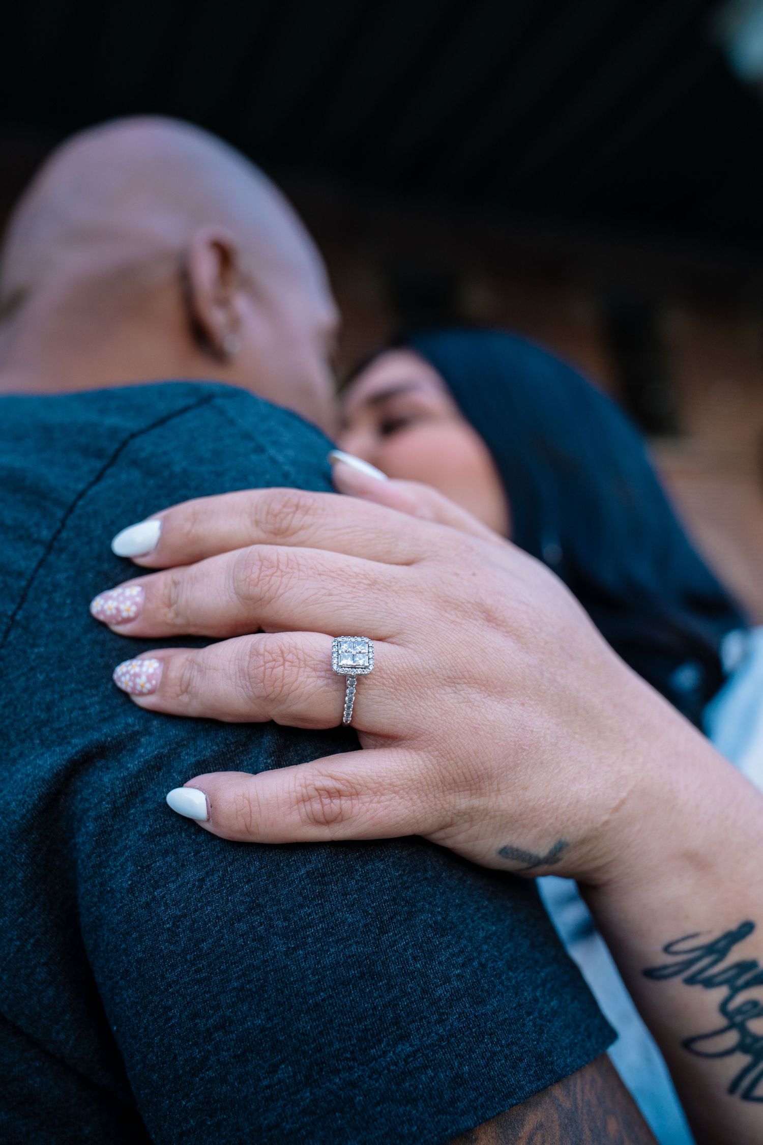 Couple embracing closely, woman's hand showing a sparkling diamond halo engagement ring with white manicured nails.