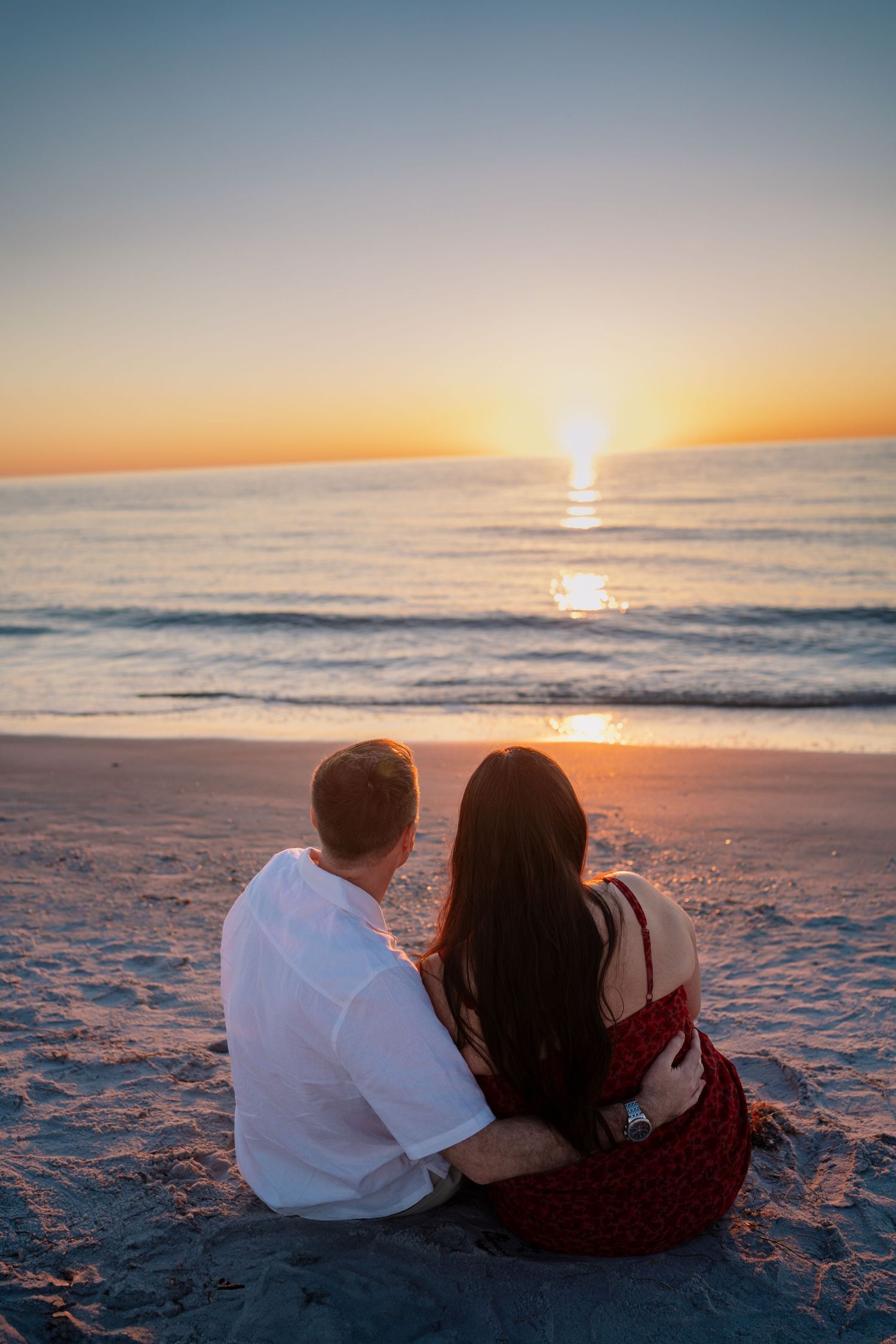 Couple sitting together on sandy beach watching a romantic golden sunset over calm ocean waves.