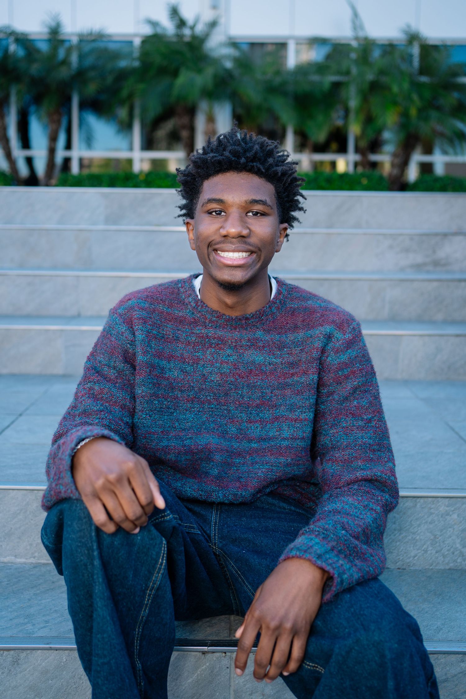 Smiling young man in striped sweater sitting on outdoor steps with palm trees in background.