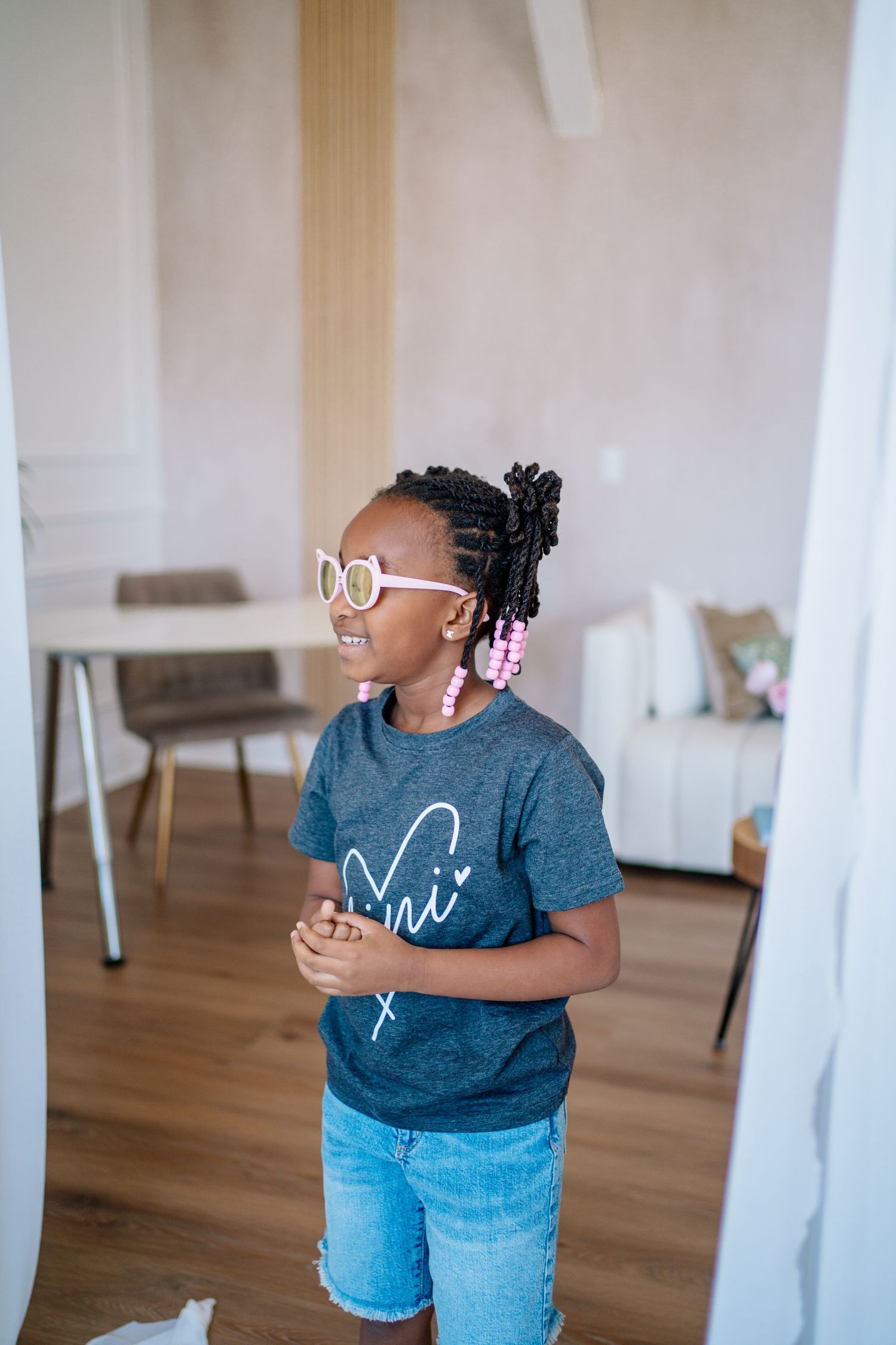 Young Black girl wearing pink sunglasses, braids, beaded earrings, and a gray t-shirt with denim shorts stands indoors.