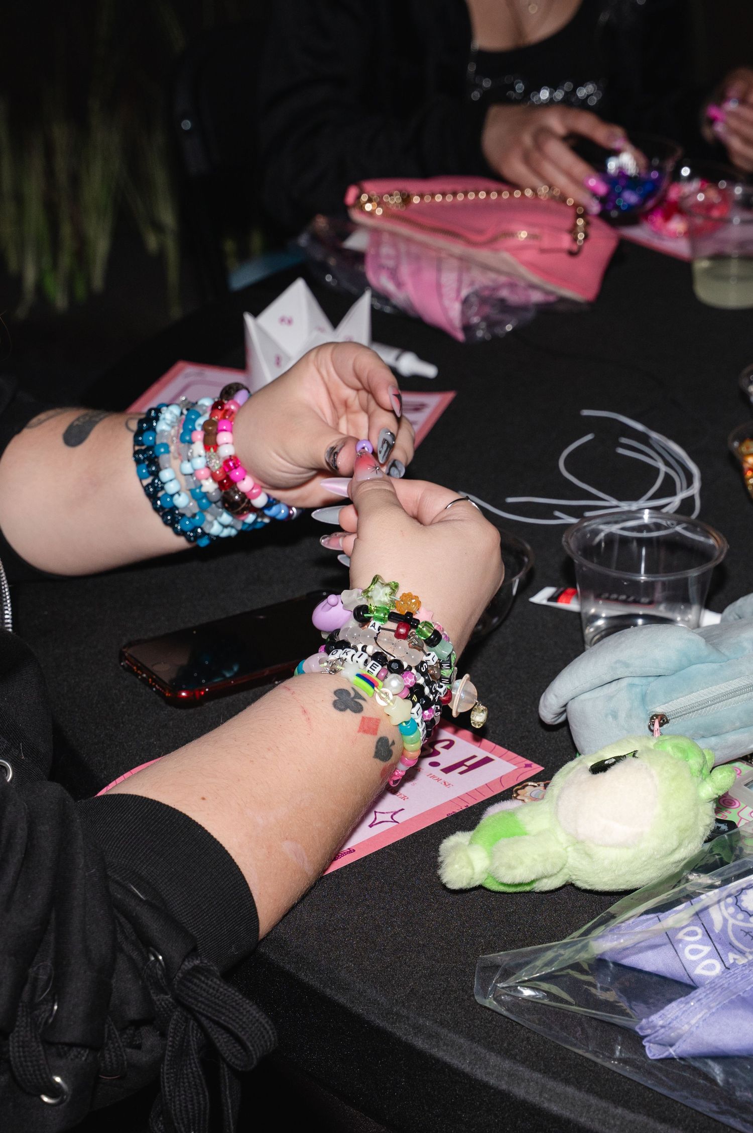Hands adorned with colorful beaded bracelets exchanging friendship bracelets at a dark table with small toys.