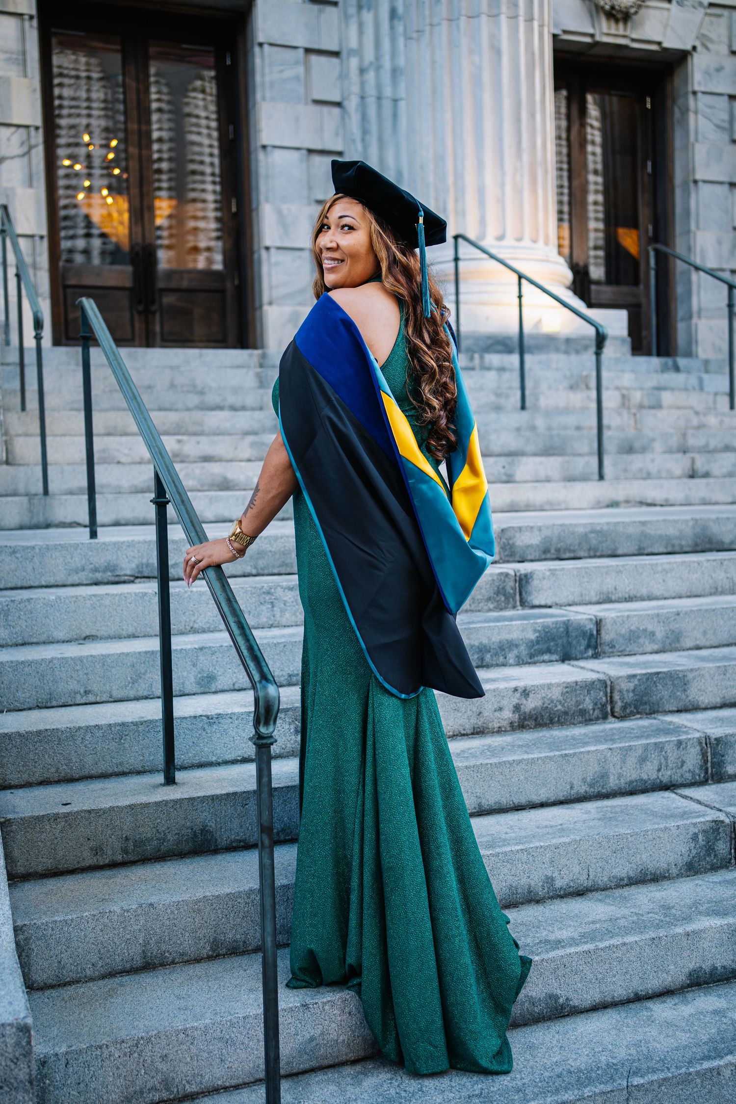Woman in green gown and graduation cap poses on stone steps, wearing blue and gold academic hood.