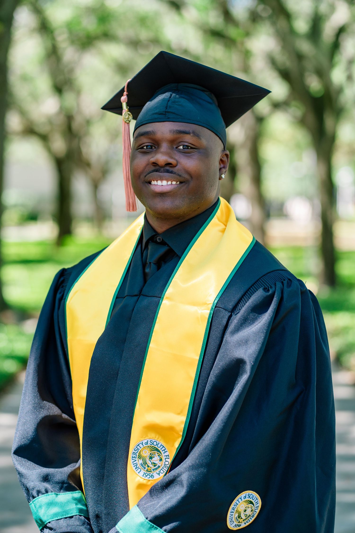 Smiling graduate in black cap and gown with gold stole posing outdoors on a sunny day.