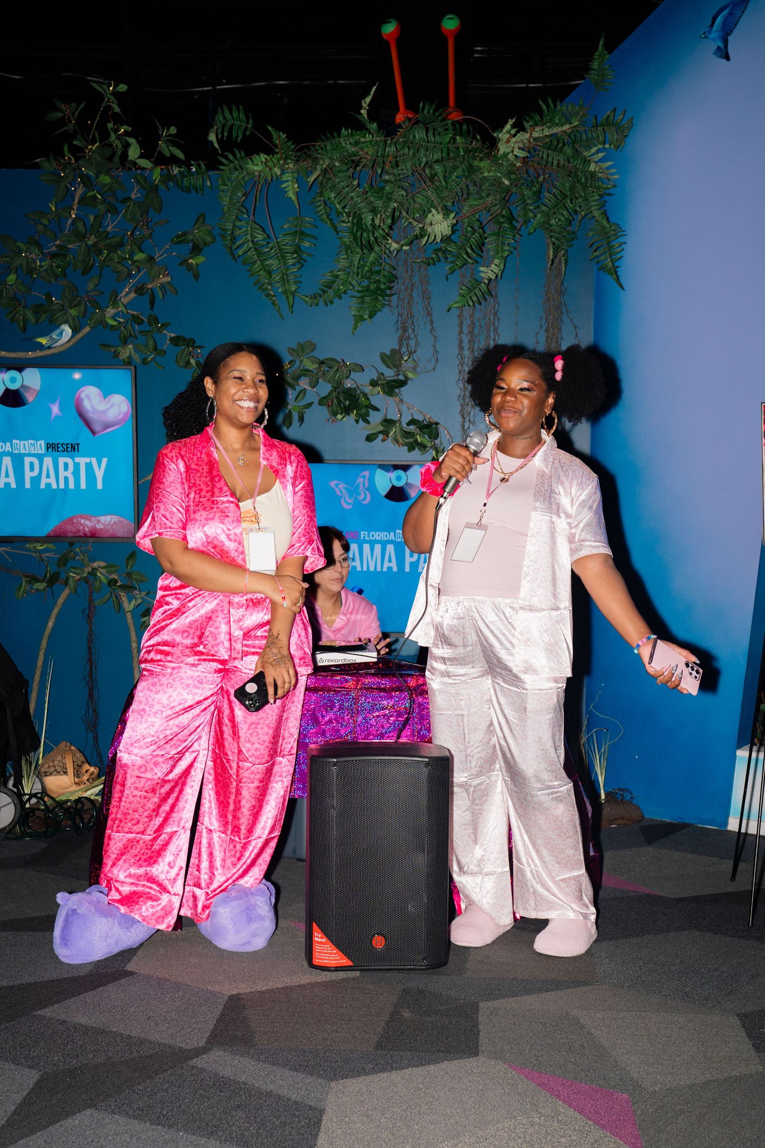 Two women in pink satin pajamas hold microphones on stage at a pyjama party event.
