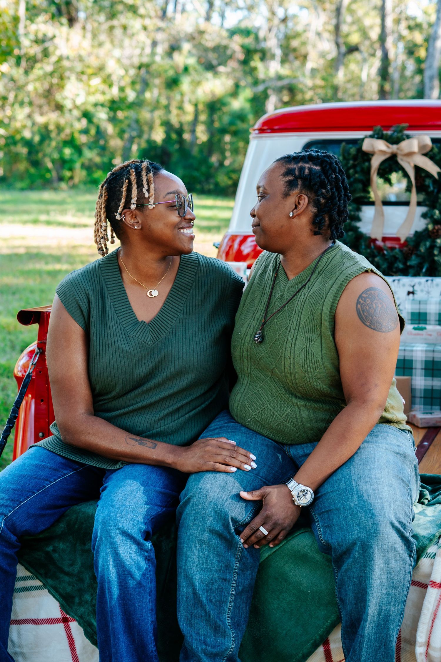 Two people in green tops and jeans sitting close together by a classic red automobile.