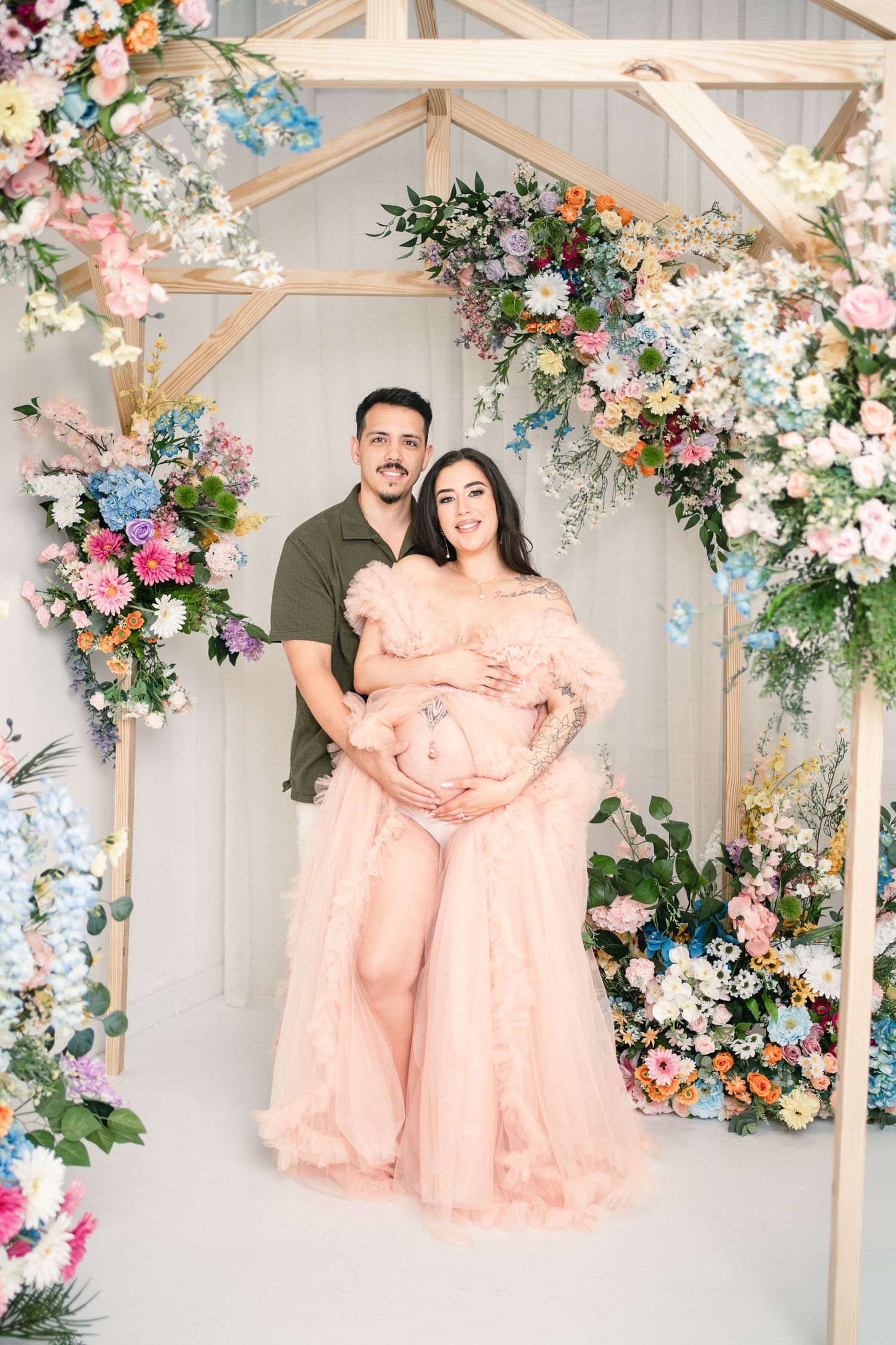 Couple poses under floral arch, pregnant woman in pink tulle gown, man in olive shirt, colorful blooms backdrop.
