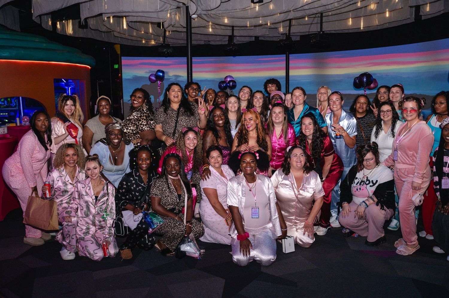 Large diverse group of women posing together in pajamas and pink outfits at a colorful indoor event venue.
