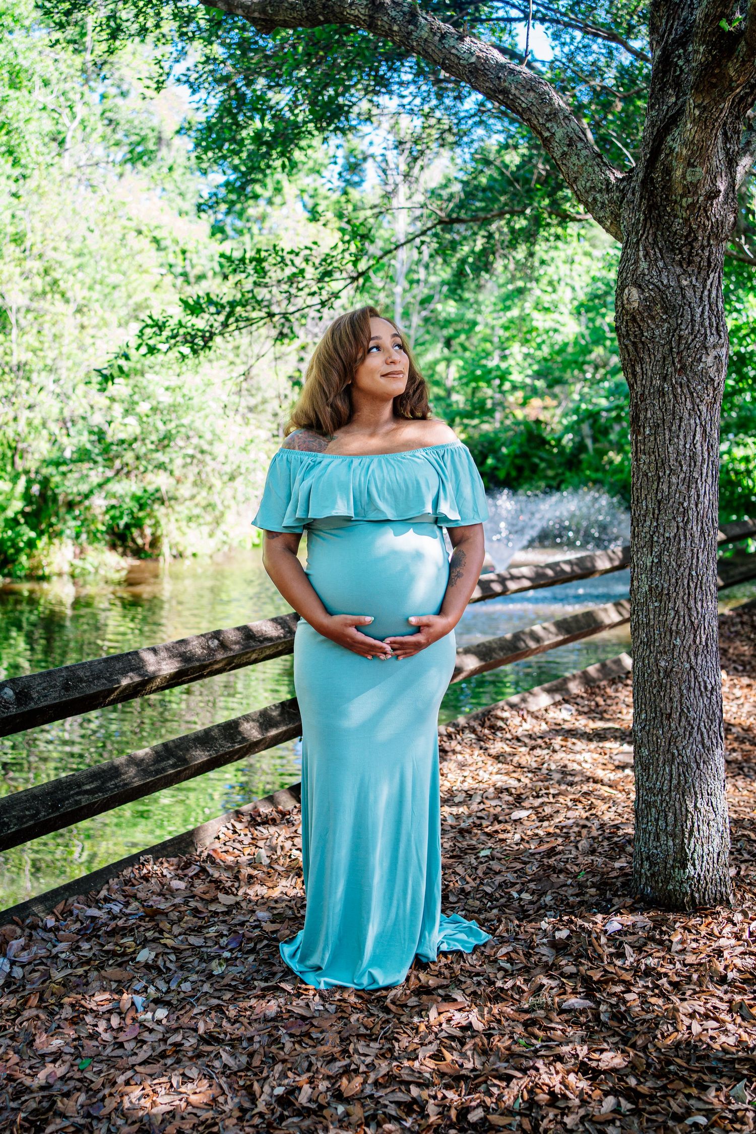 Pregnant woman in teal off-shoulder gown cradling belly outdoors near a serene pond surrounded by lush trees.