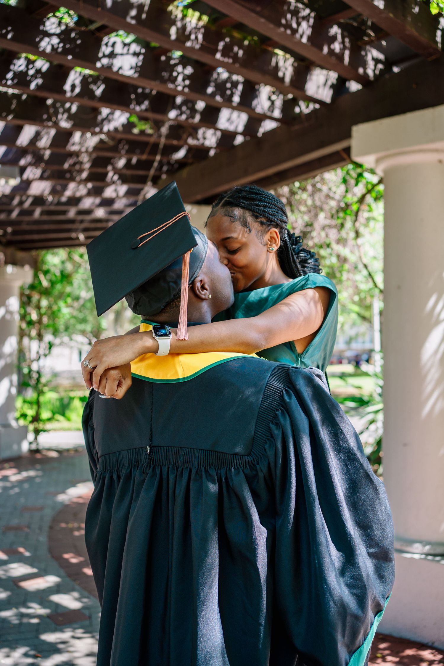 Two graduates in green caps and gowns share a warm embrace during a graduation celebration outdoors.