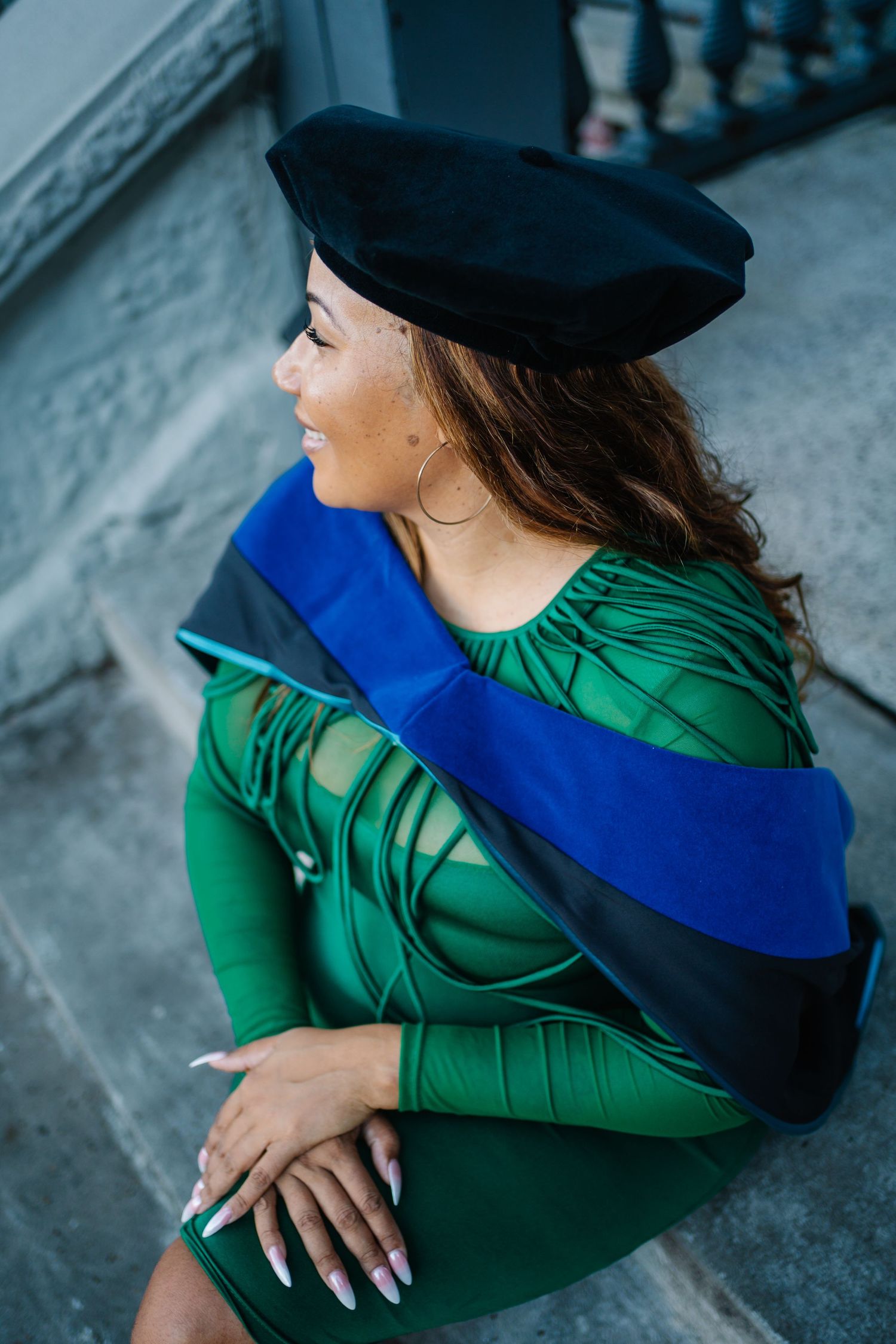 Woman in green dress wearing black graduation beret and blue academic hood, sitting on steps, smiling.