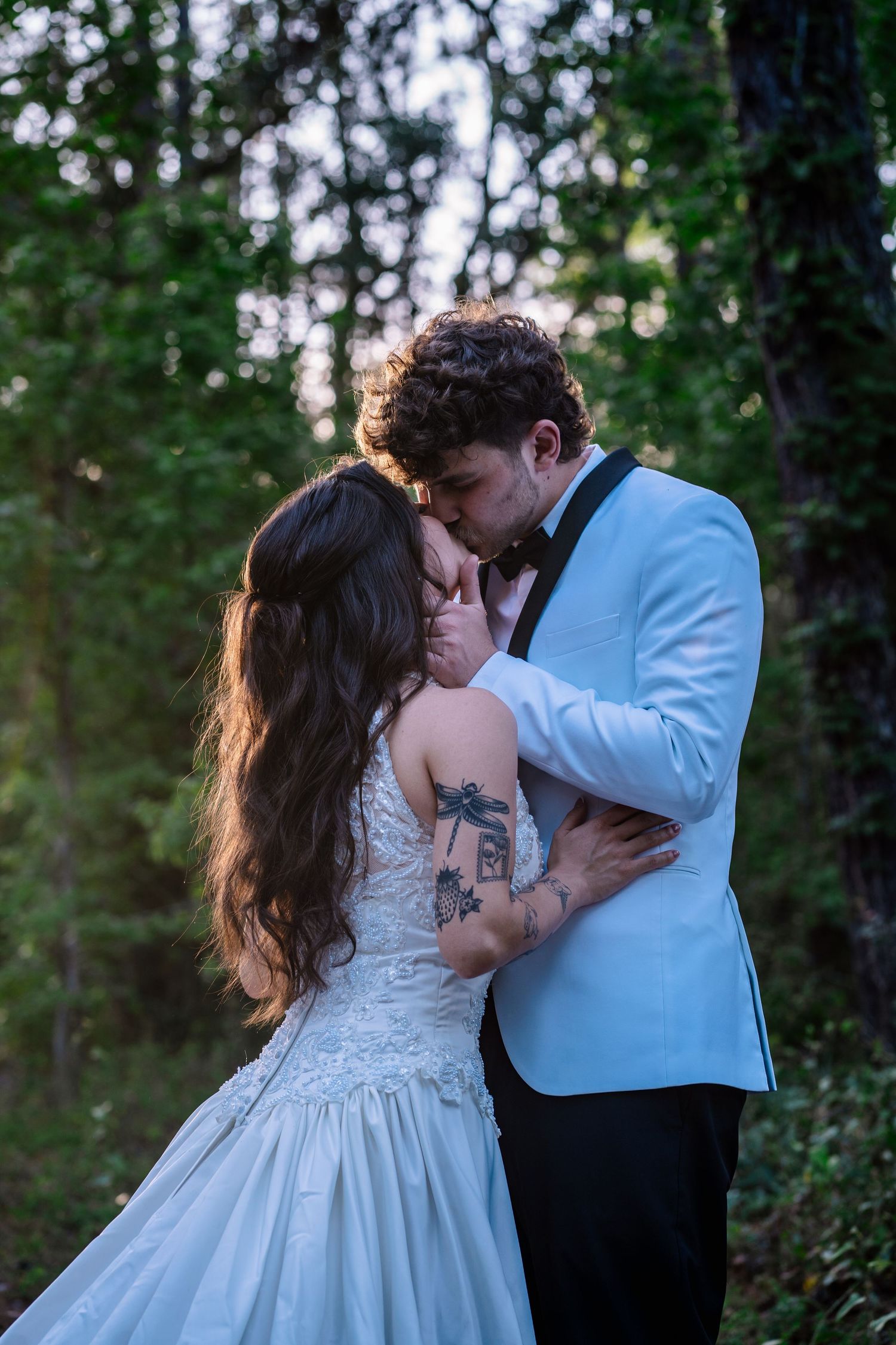 A bride and groom share a romantic kiss in a lush forest setting, with the bride wearing a lace gown and showing tattoos.