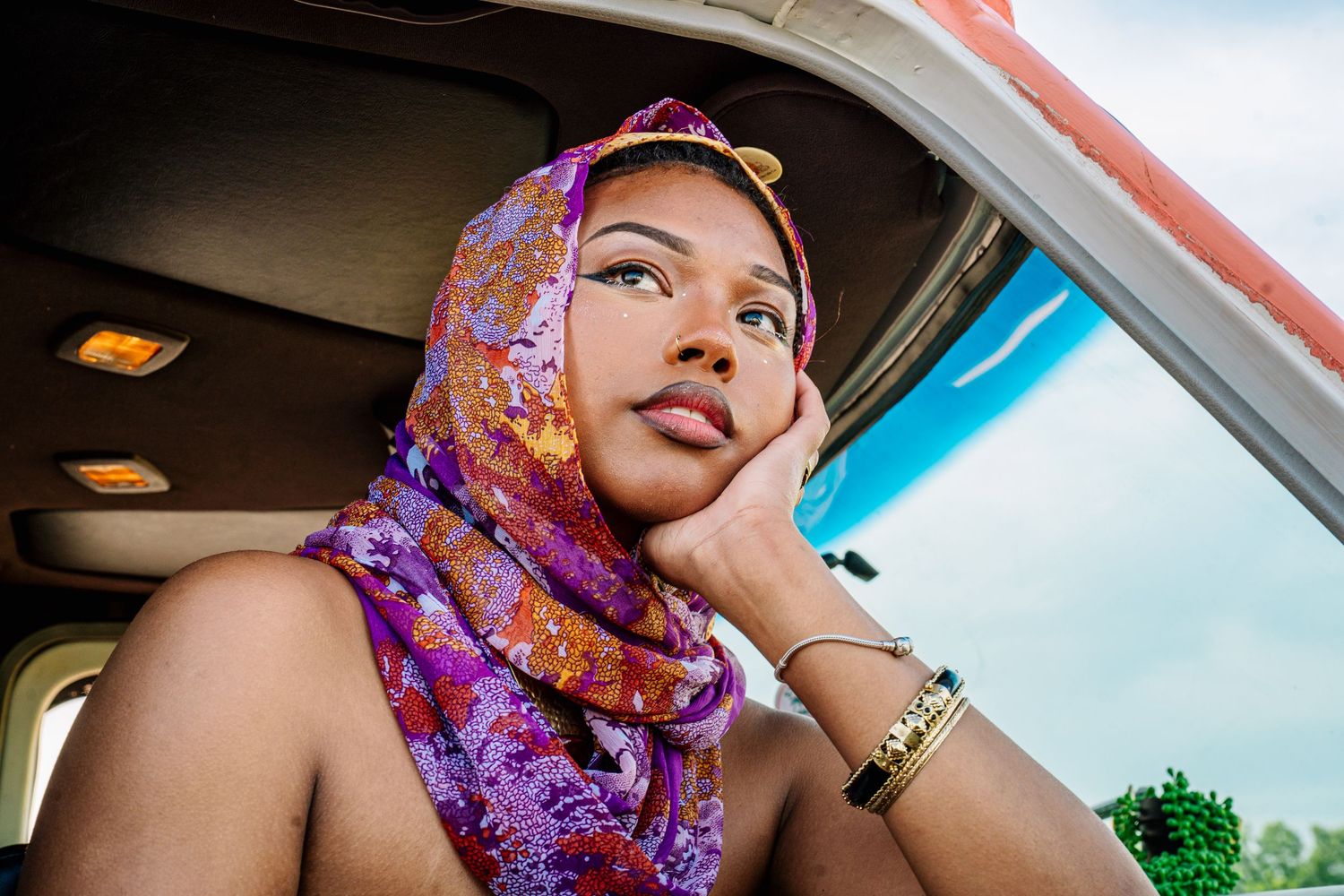 Young woman wearing colorful purple floral hijab sitting in a vehicle, resting chin on hand.