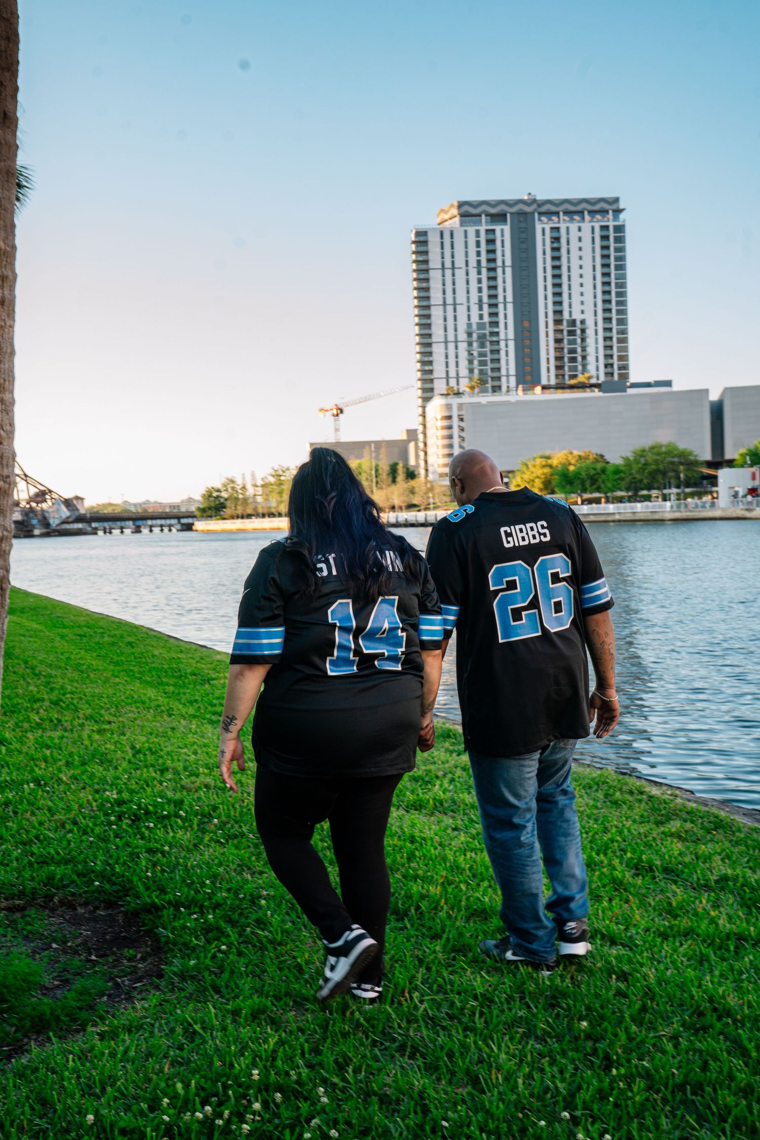 Couple walking by waterfront in matching black Gibbs #26 and #14 NFL jerseys on green grass with city skyline.