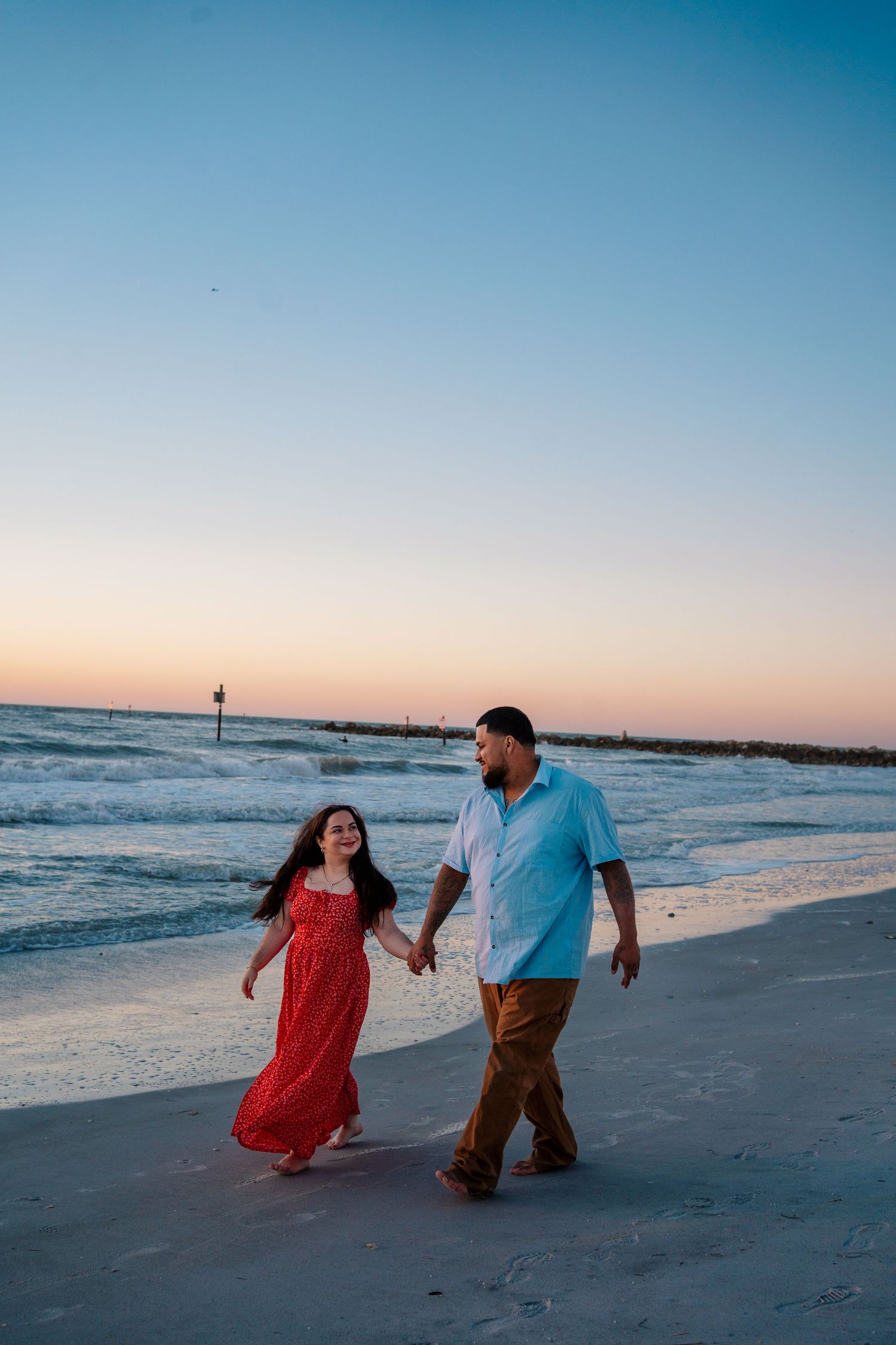 Couple holding hands walking on beach at sunset, woman in red dress, man in blue shirt, ocean waves behind them.