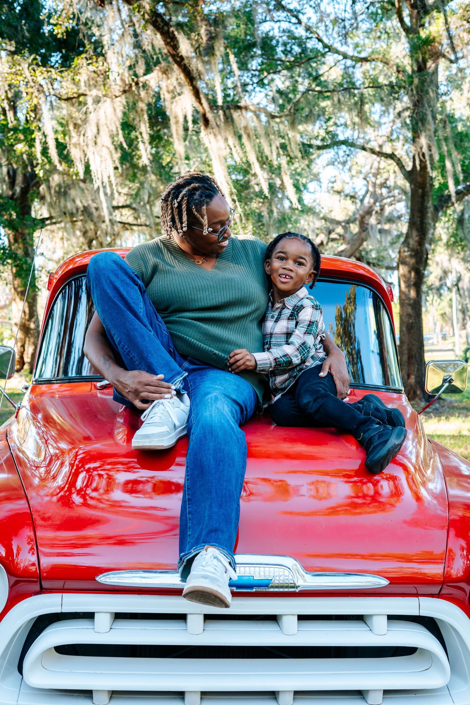 A couple shares a playful moment while lying on the hood of a bright red vintage car in a wooded setting.