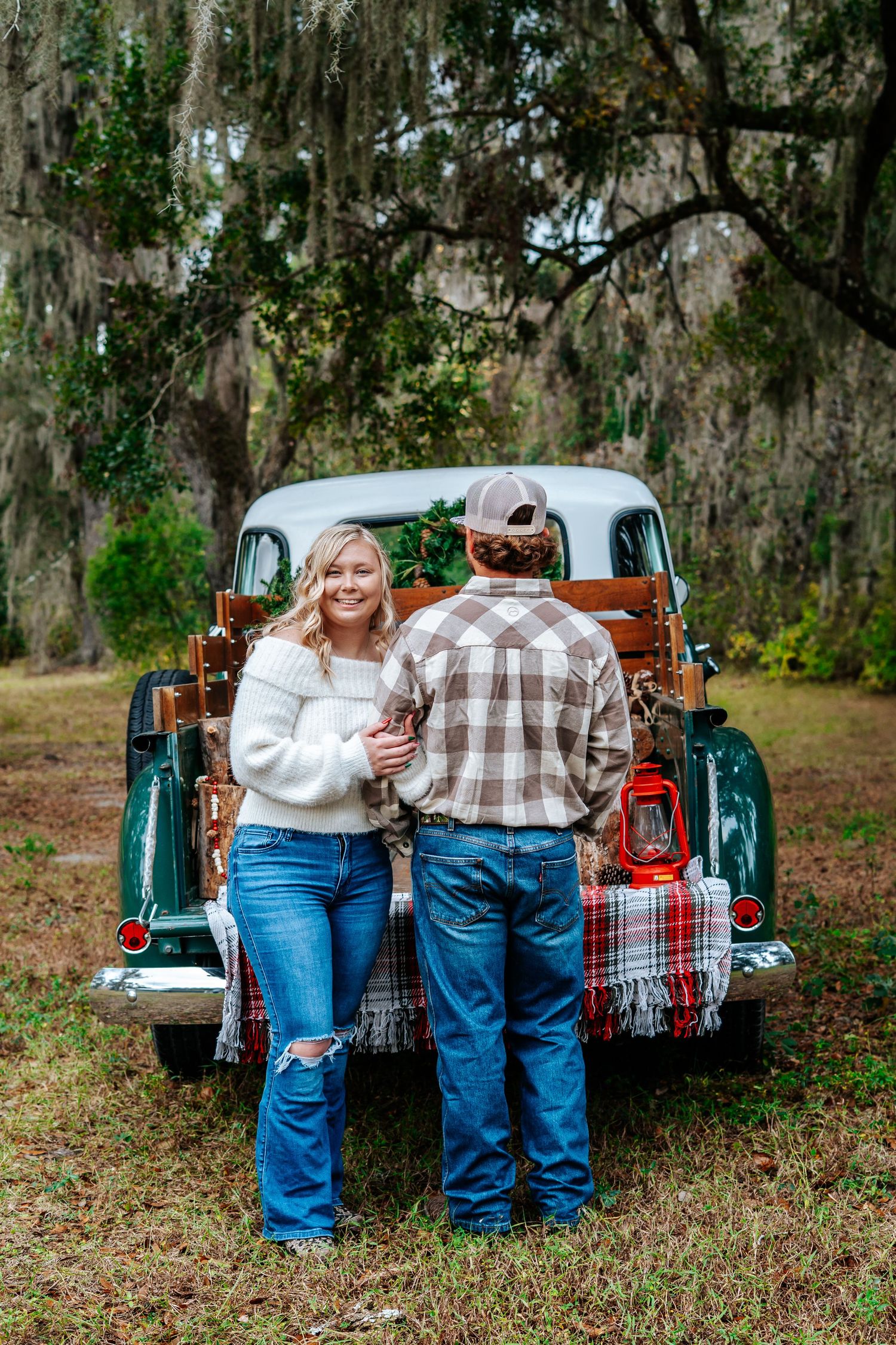 Casual couple posing by classic pickup truck with plaid blanket in wooded setting.