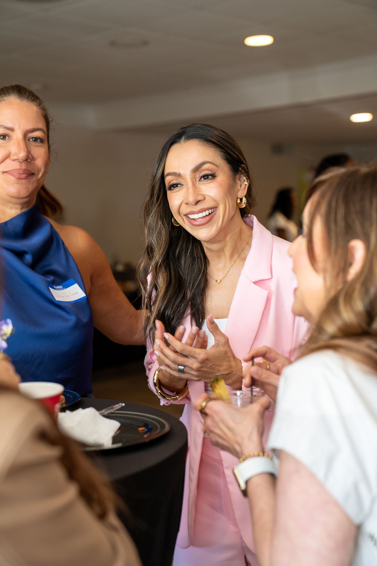 Women networking at a social event, smiling and conversing in a bright venue, one wearing a pink blazer.