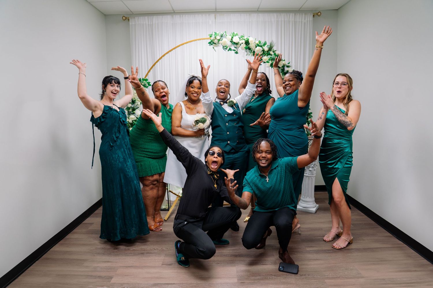 Joyful wedding party in emerald green dresses celebrating near a floral hoop arch with hands raised.