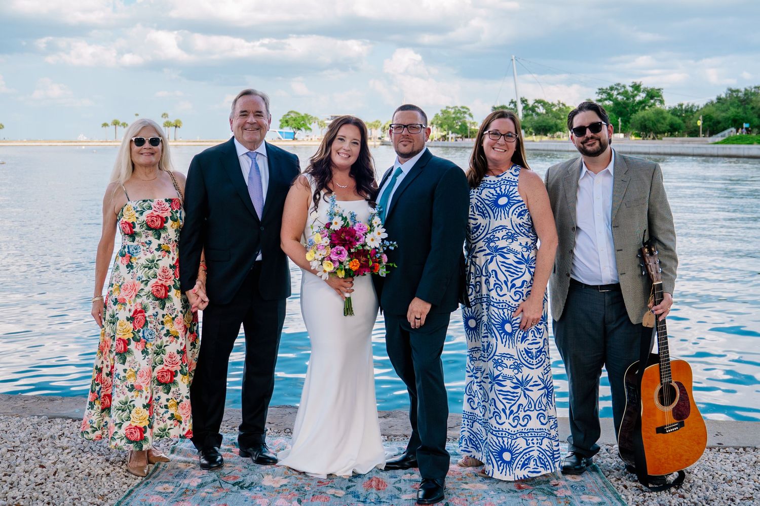 Wedding party poses outdoors near waterfront, bride in white gown holding bouquet, guests in formal attire, guitarist nearby.