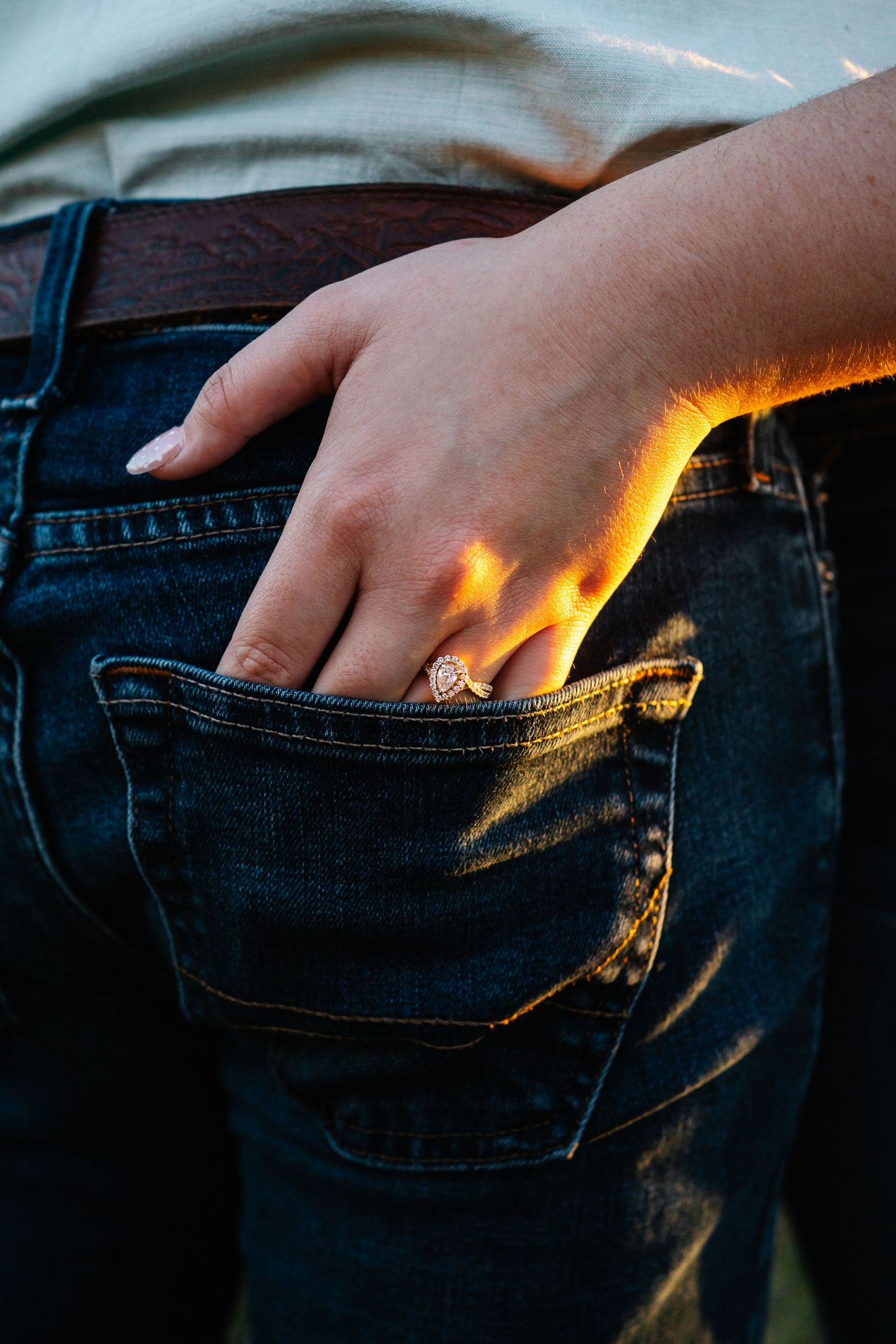 Close-up of a hand with a gold ring tucked into a back pocket of dark blue jeans, lit by warm golden sunlight.