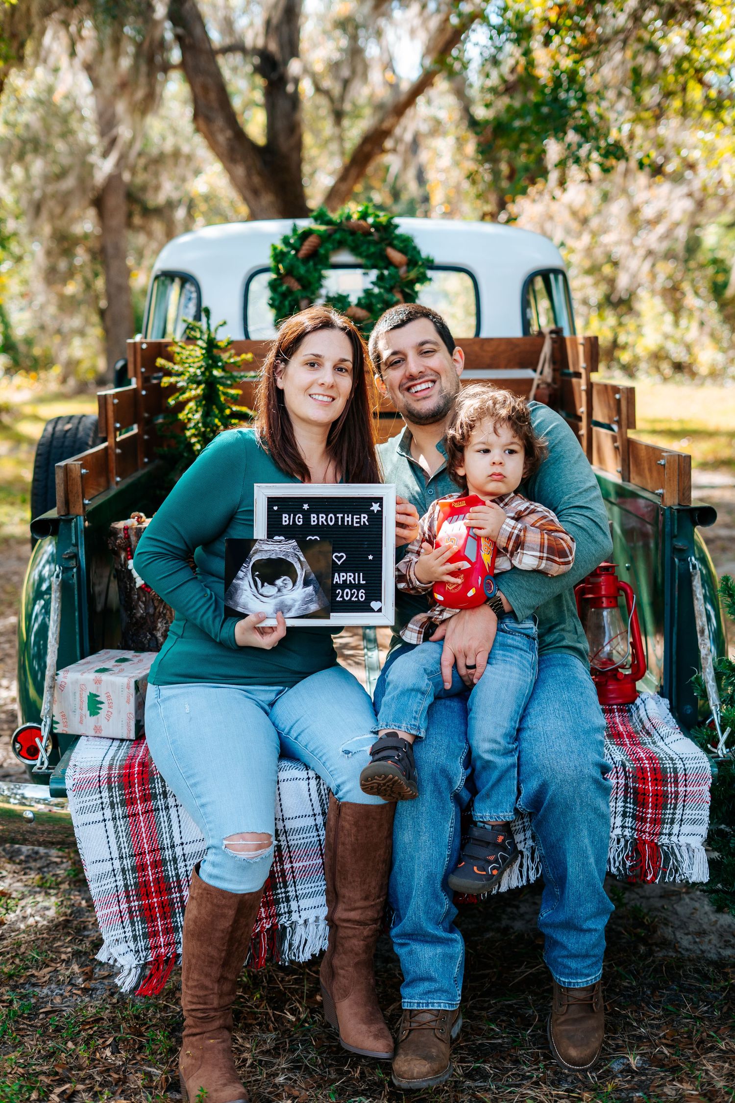 Family pregnancy announcement photo with couple and toddler holding Big Brother sign by vintage truck decorated for Christmas.