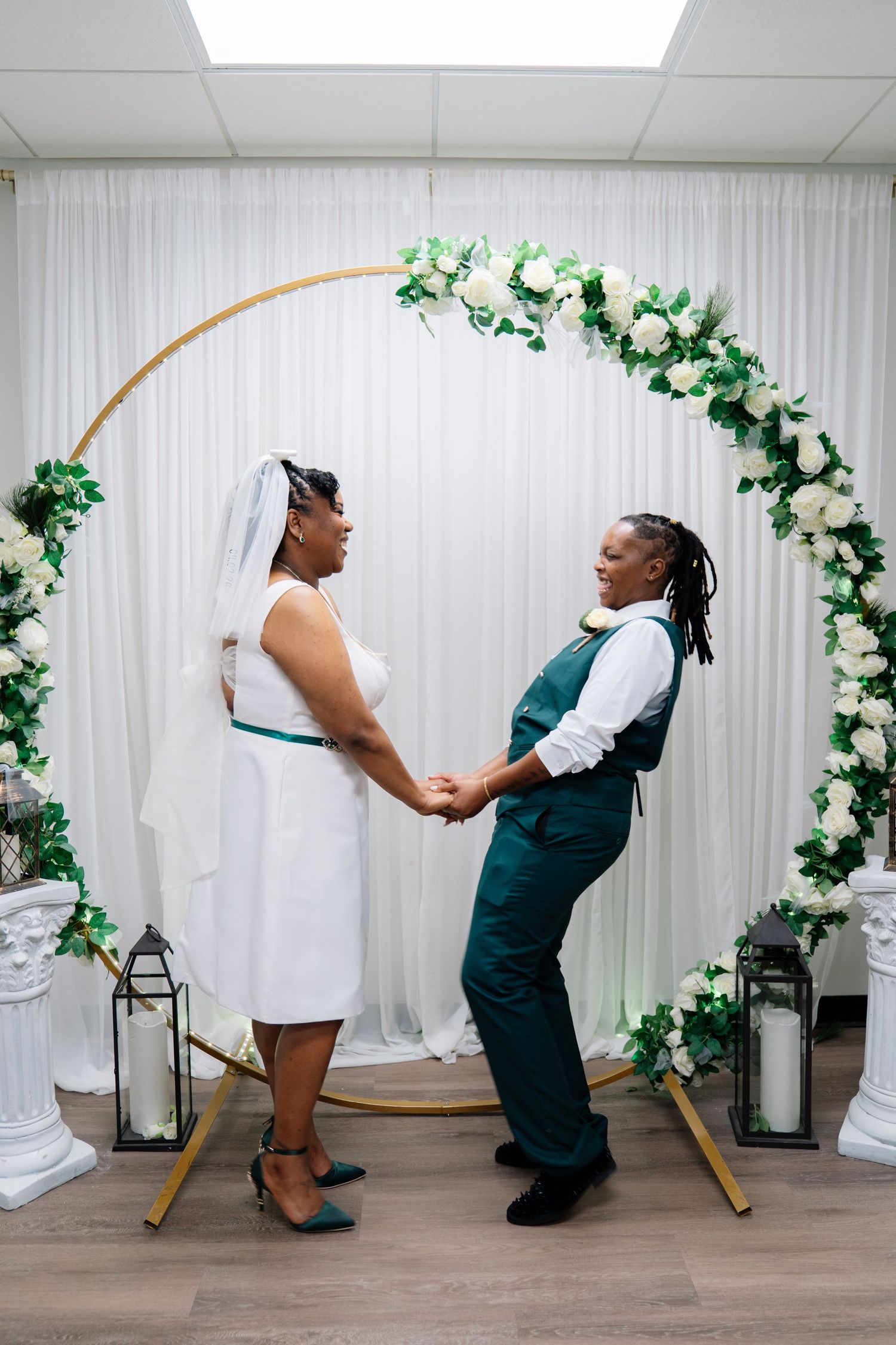 Two brides hold hands at wedding altar with circular floral arch decorated with white roses and greenery.