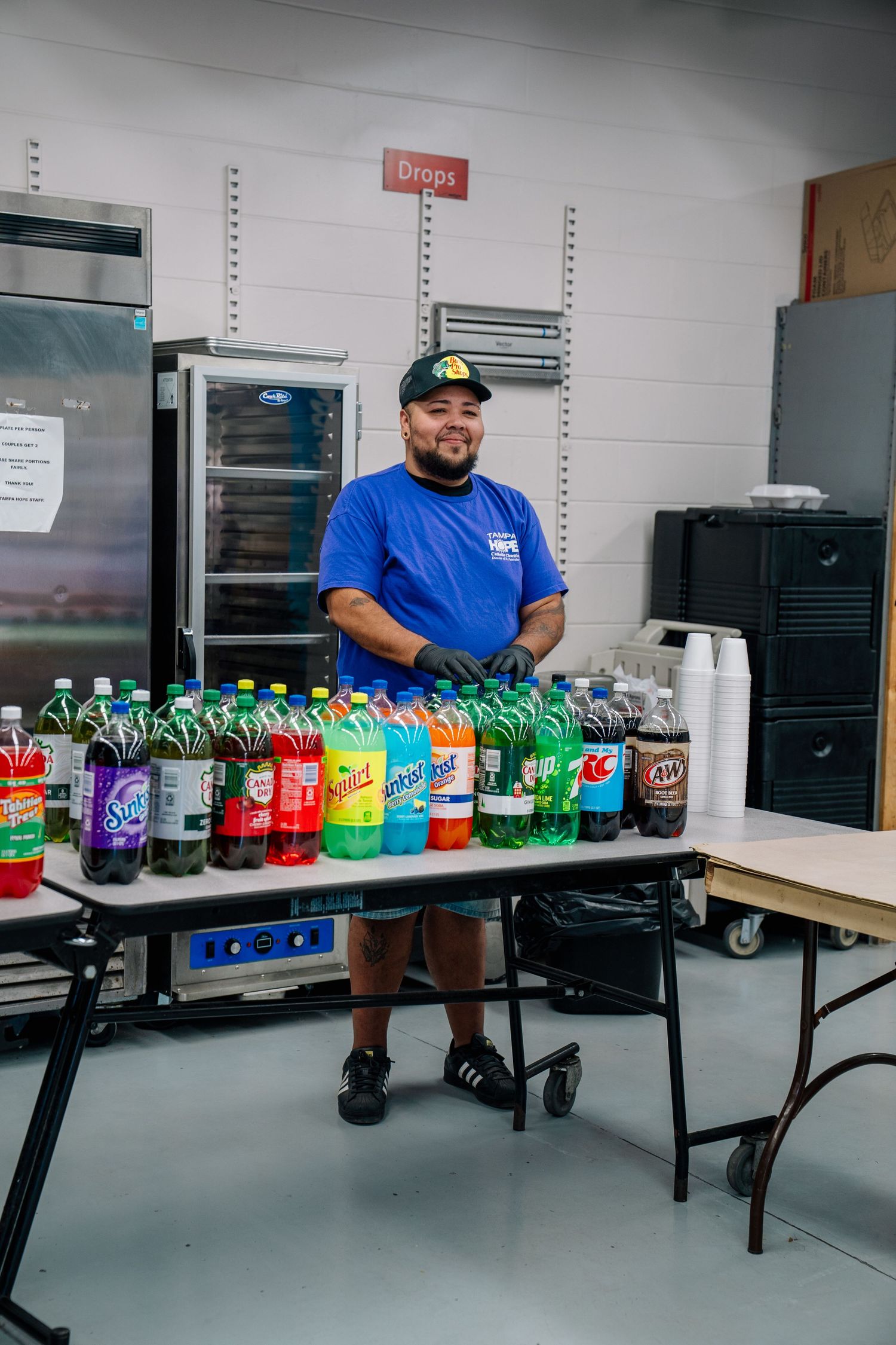 Smiling man in blue shirt stands behind table of assorted soda bottles at a community food distribution event.