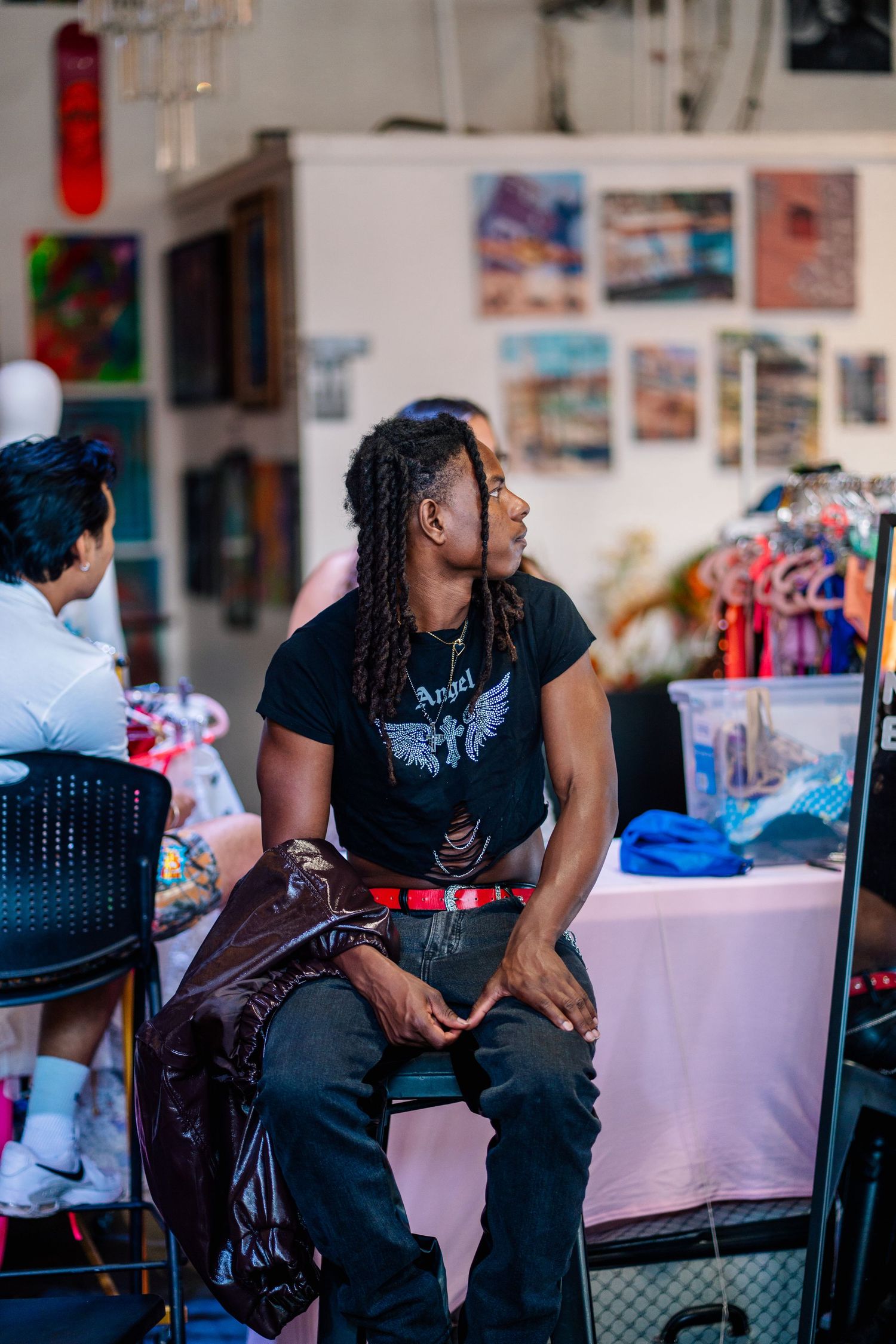 Young man with dreadlocks wearing a black Angel graphic tee sits in an art studio surrounded by colorful artwork.