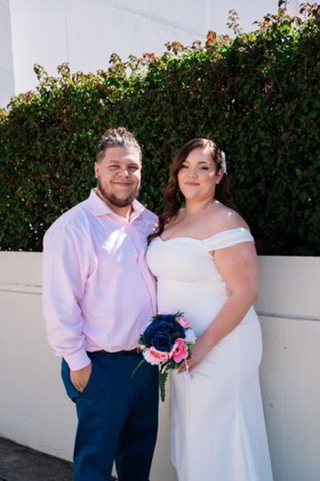 Wedding couple in pink shirt and white dress pose together in front of a garden wall outdoors.