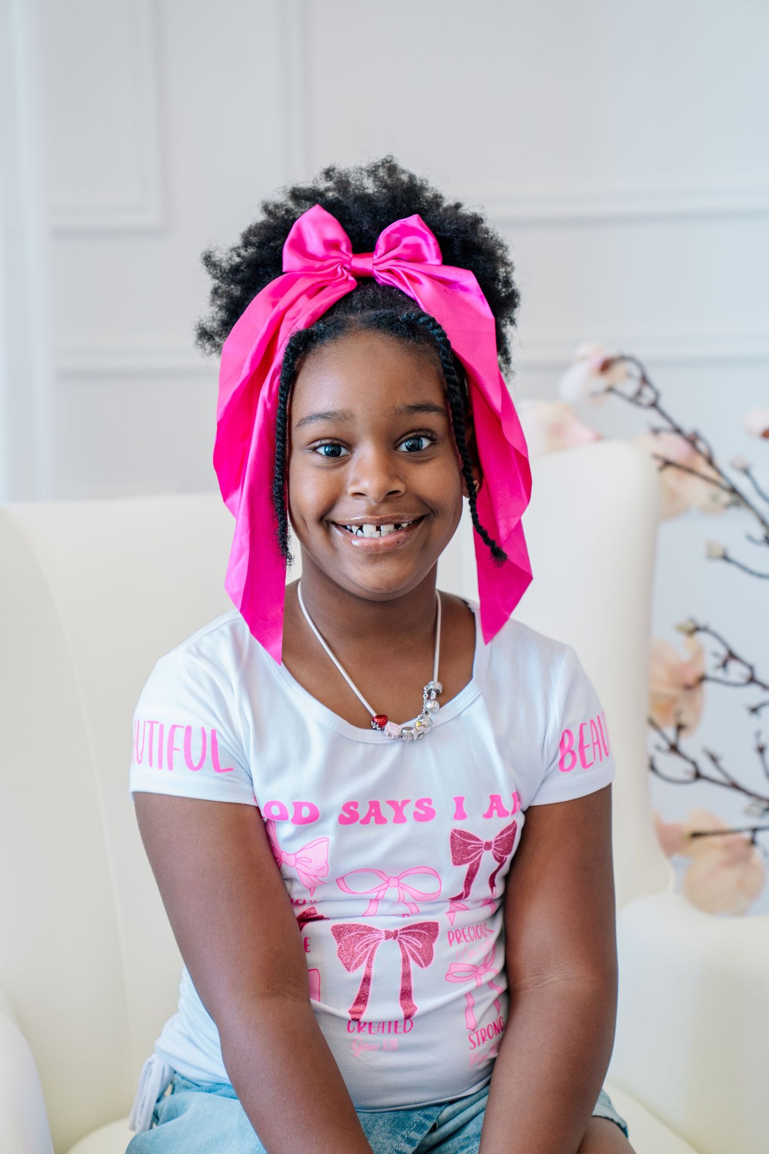 Smiling young girl with curly hair wearing a pink bow headband and white graphic t-shirt, sitting on a cream chair.