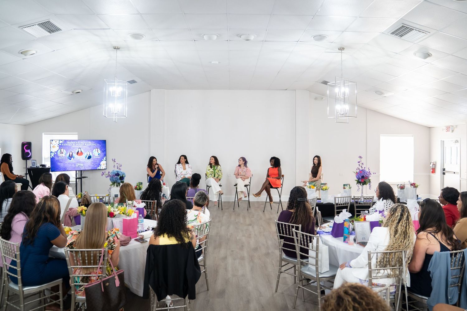 Women gathered in a bright event hall for a panel discussion, seated at decorated tables facing speakers on a small stage.