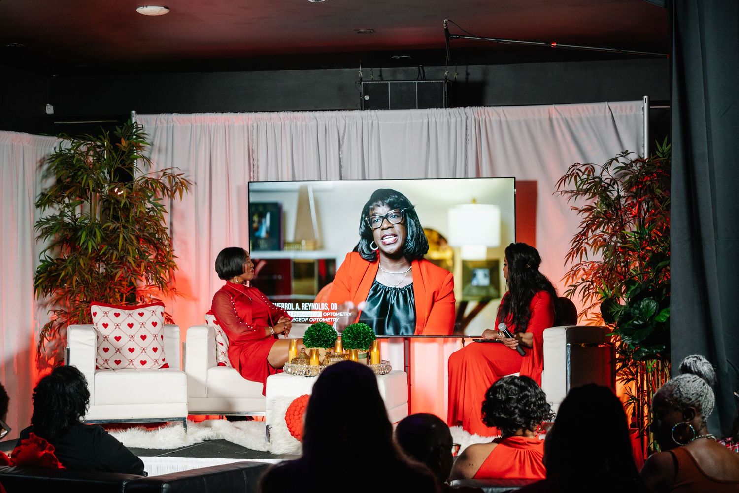 Panel discussion on stage with speakers in red outfits and a virtual presenter on screen at a women empowerment event.