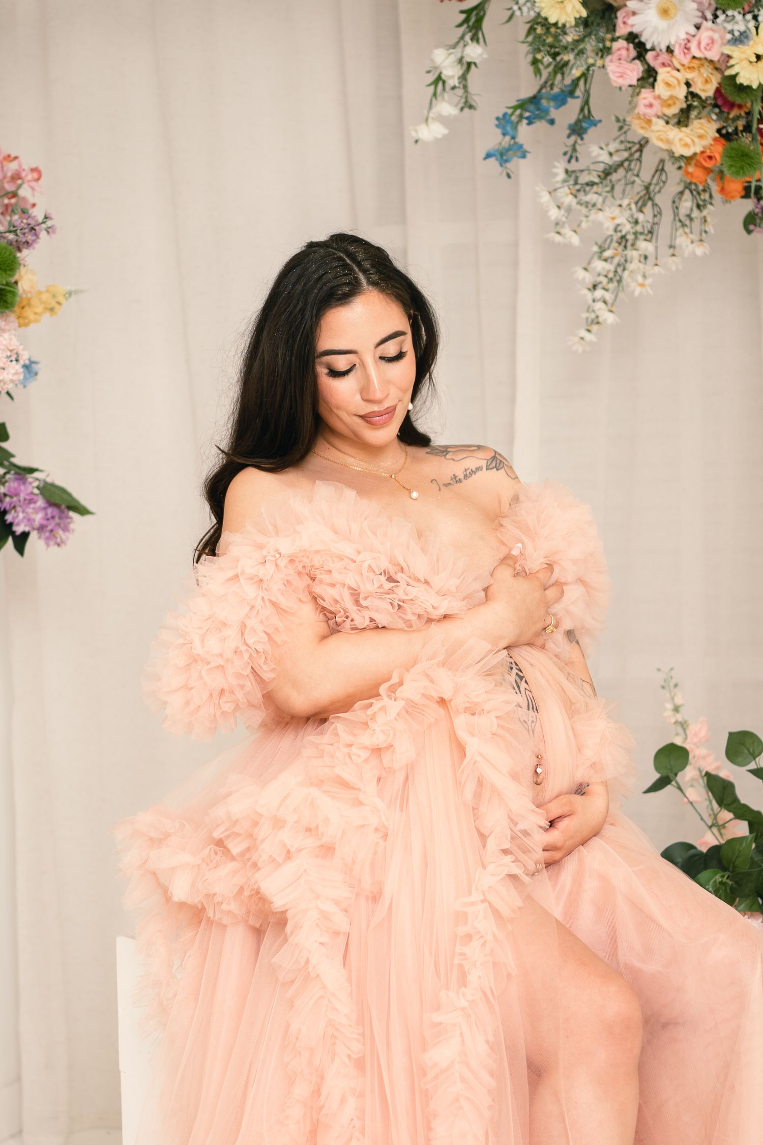 Pregnant woman in elegant blush pink ruffled off-shoulder gown poses gracefully surrounded by colorful floral arrangements.