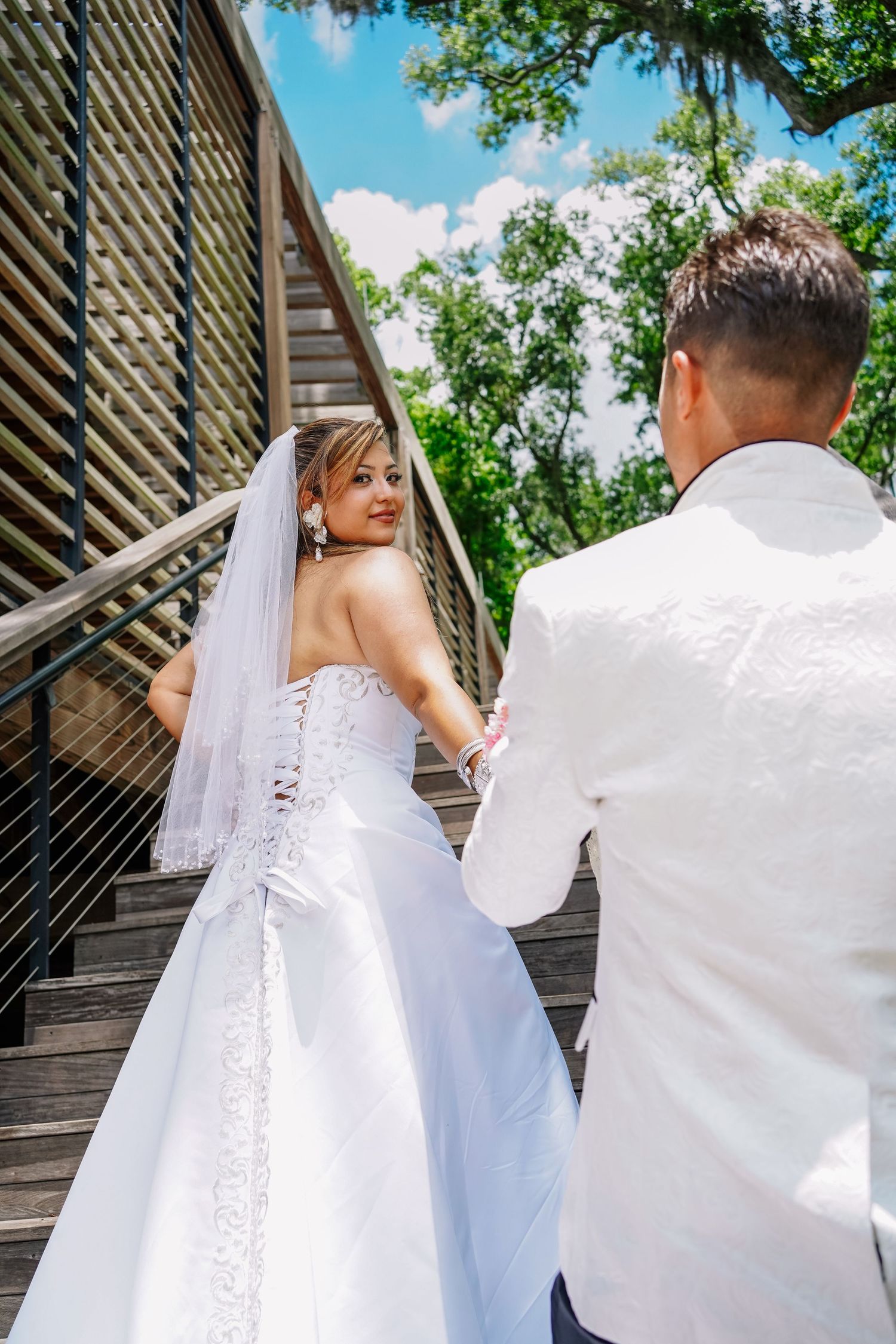 Bride in white gown and veil holds groom's hand on wooden bridge stairs, glancing back, surrounded by green trees.