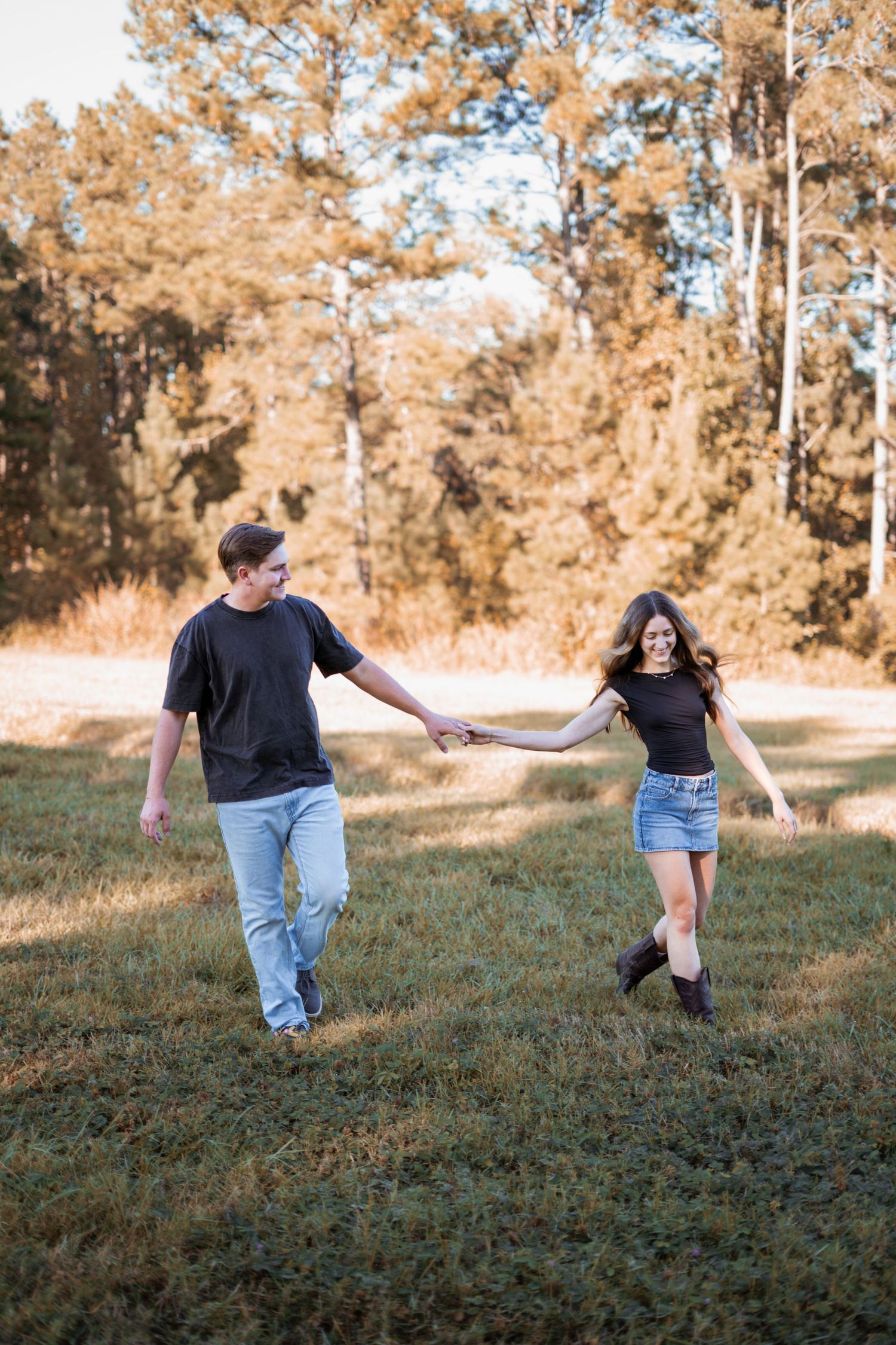 A couple running and laughing together in a field with pine trees during sunset.