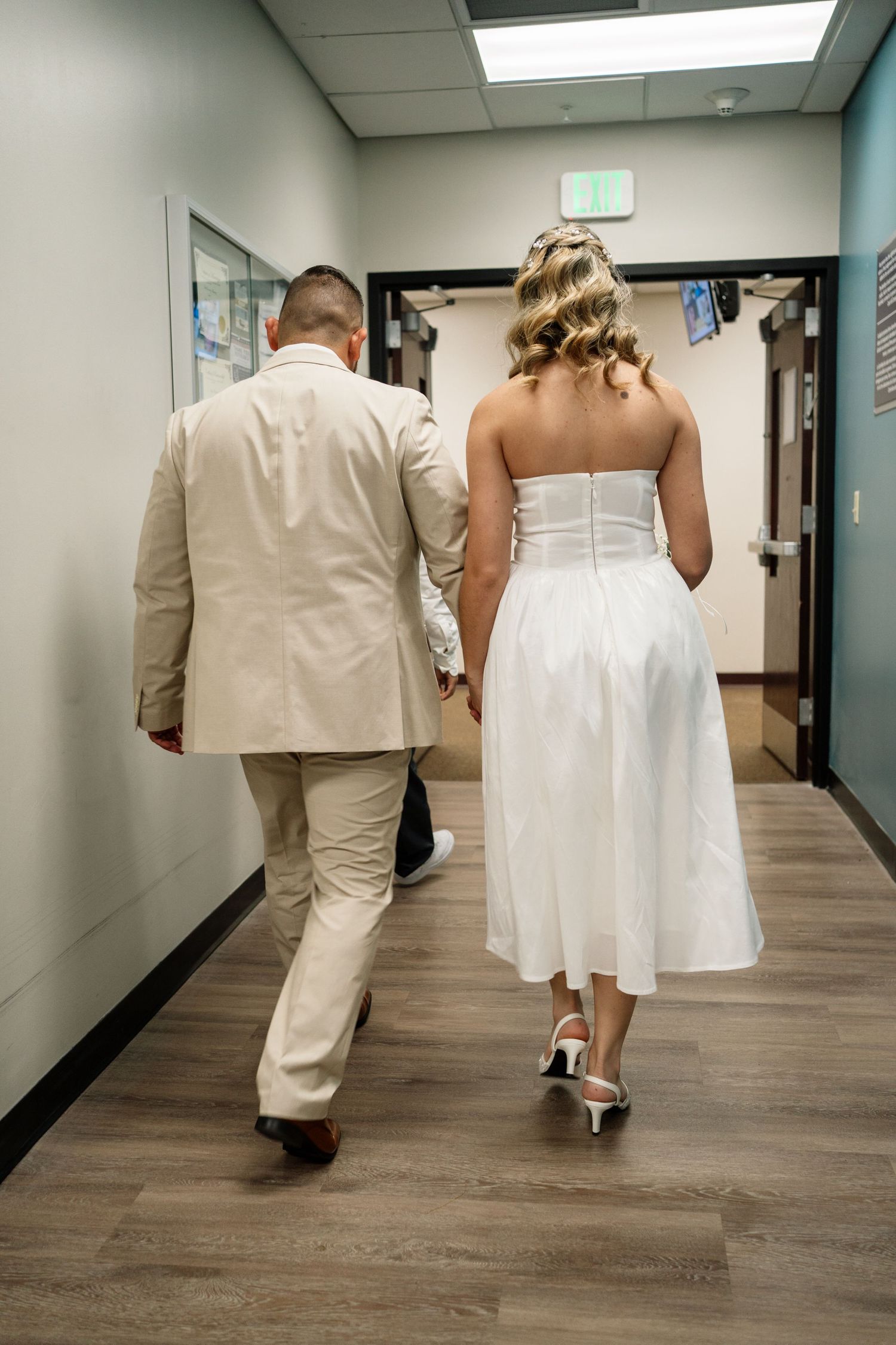 A couple in wedding attire walks down a modern hallway with wood floors and white walls.
