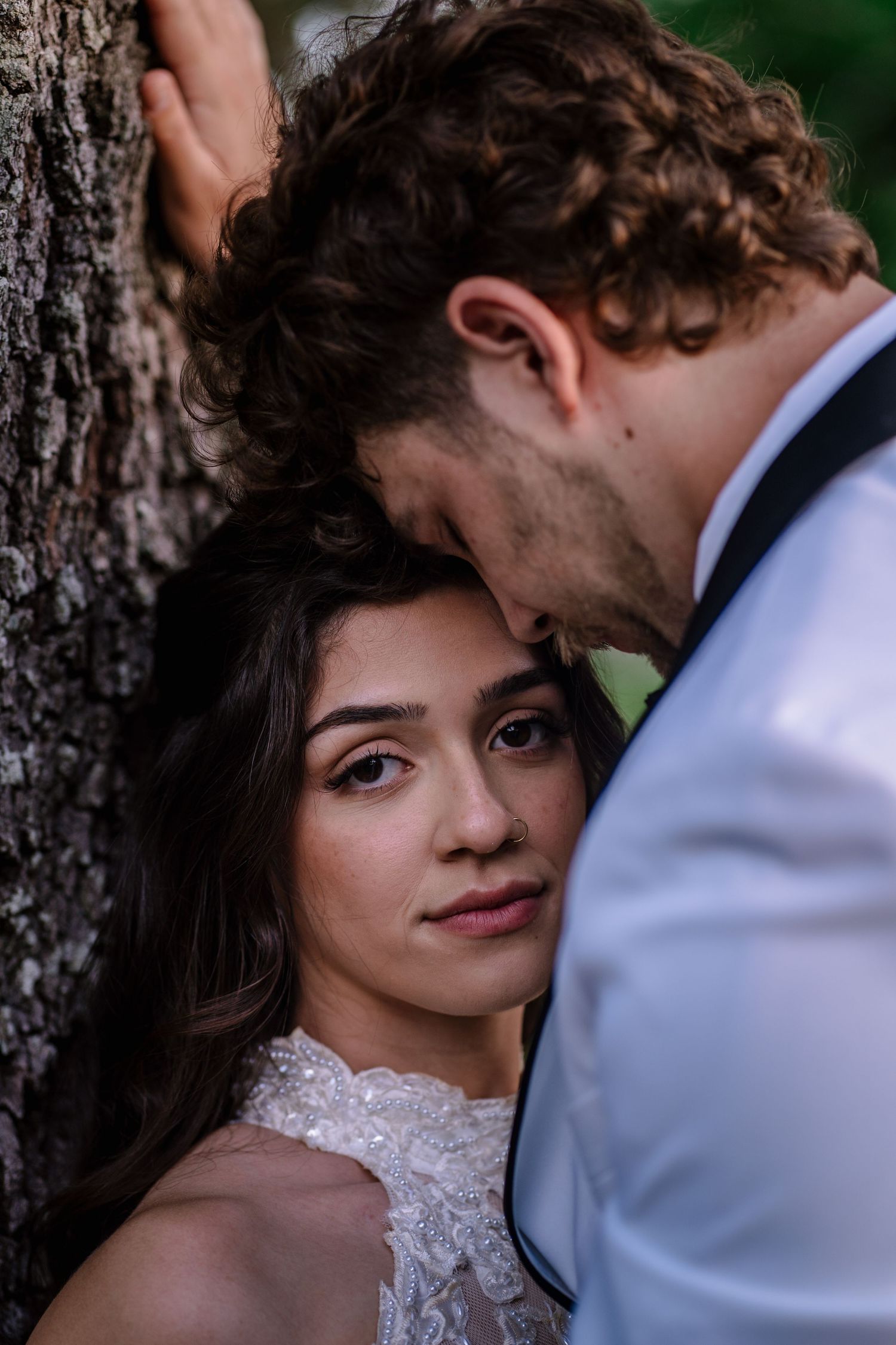 A bride in a lace gown leans against a tree beside a groom in a white suit, sharing an intimate moment outdoors.