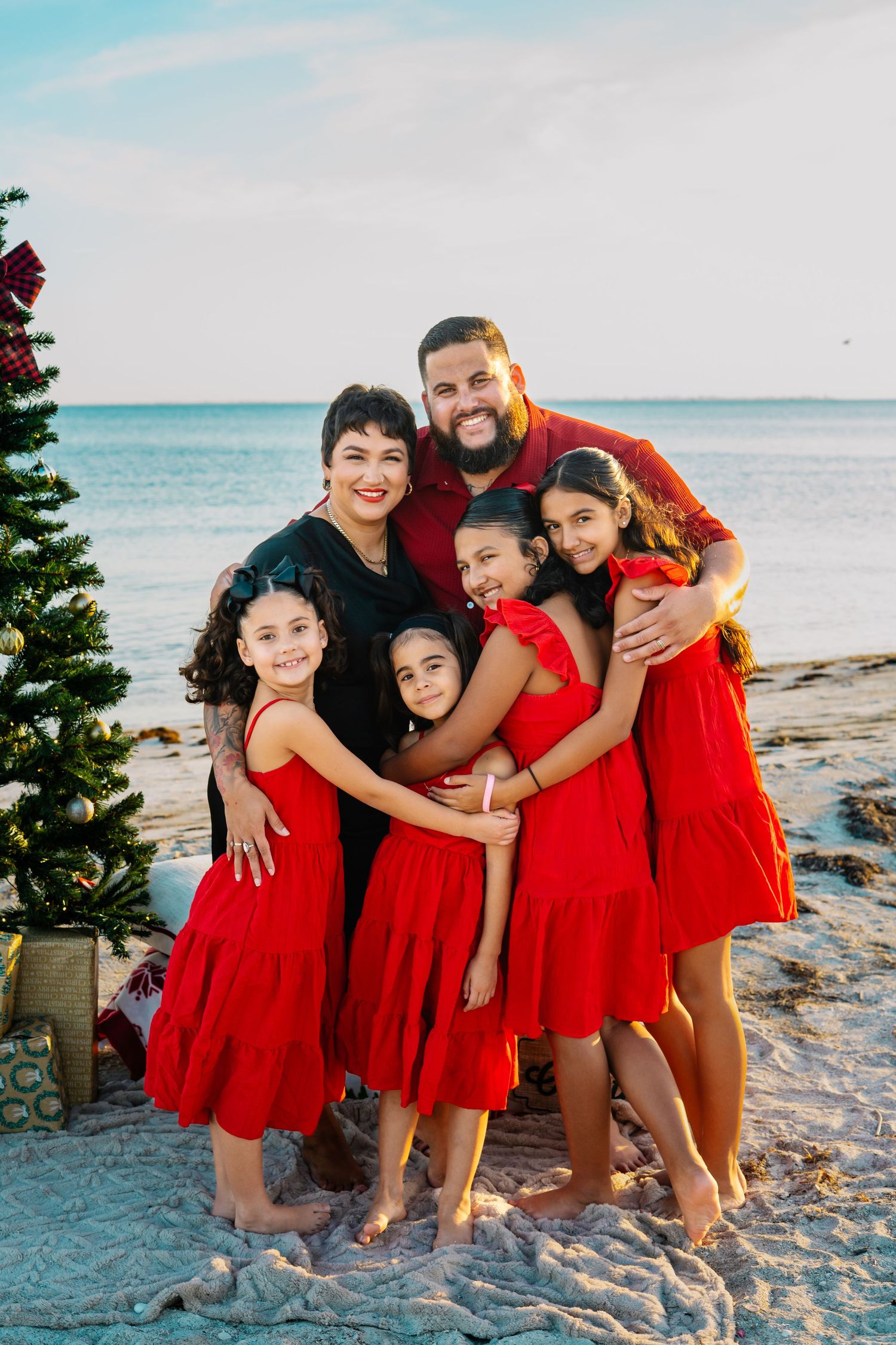 Family of five posing together on a beach beside a Christmas tree, wearing red outfits, smiling warmly at sunset.