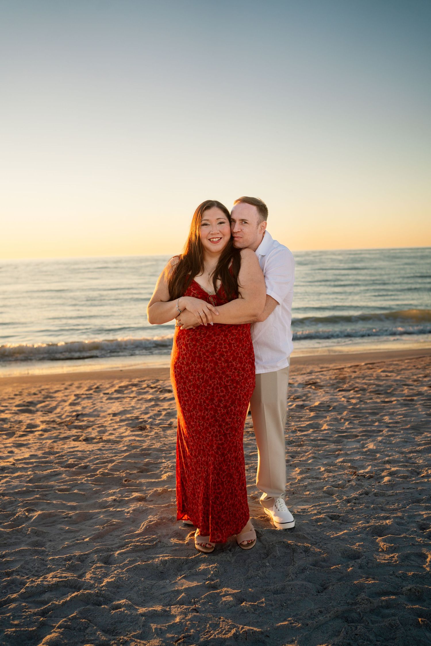 Couple embracing on a sandy beach at sunset, woman in red floral dress, man in white shirt, ocean in background.