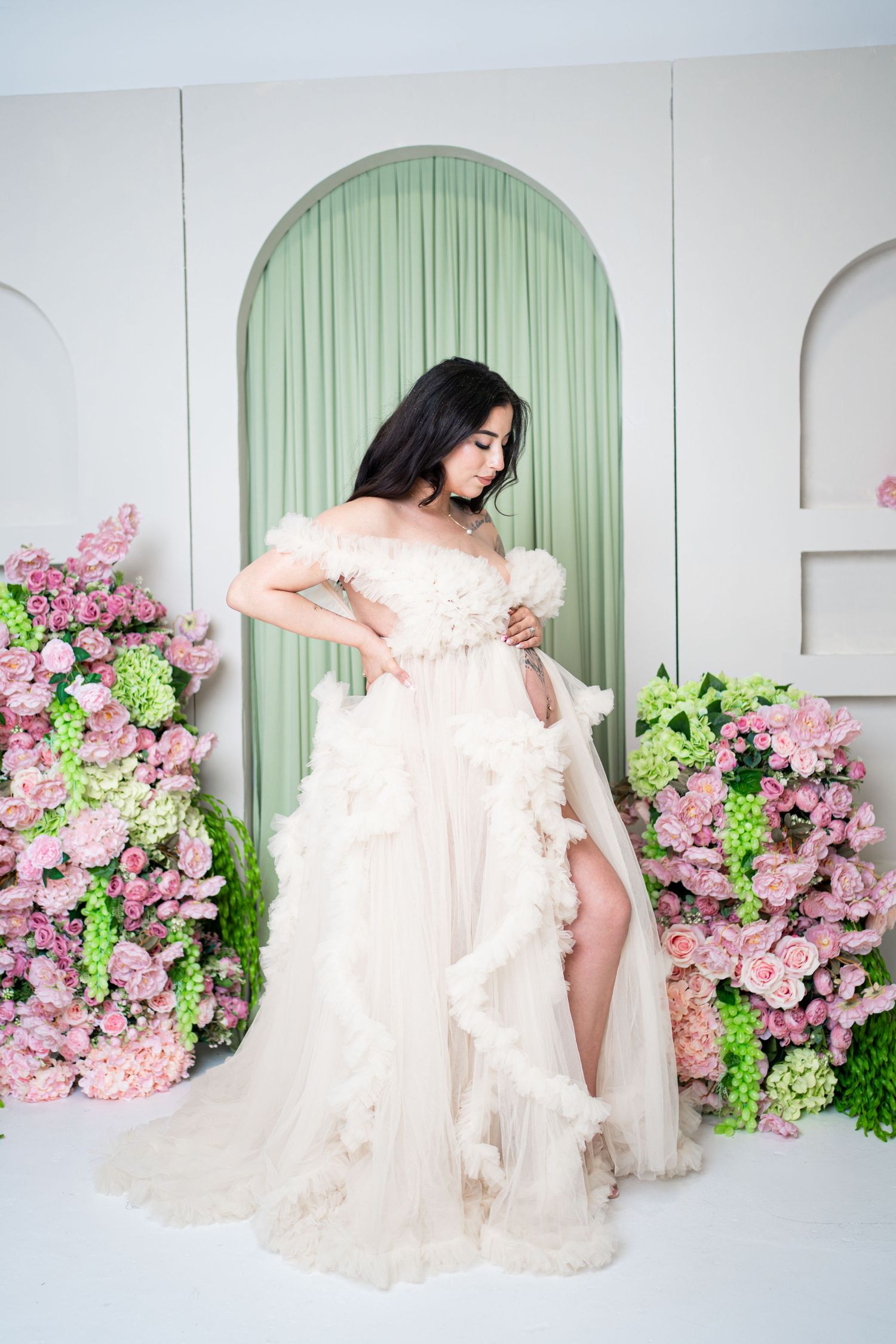 Pregnant woman in white ruffled off-shoulder gown poses gracefully among pink floral arrangements in elegant studio.